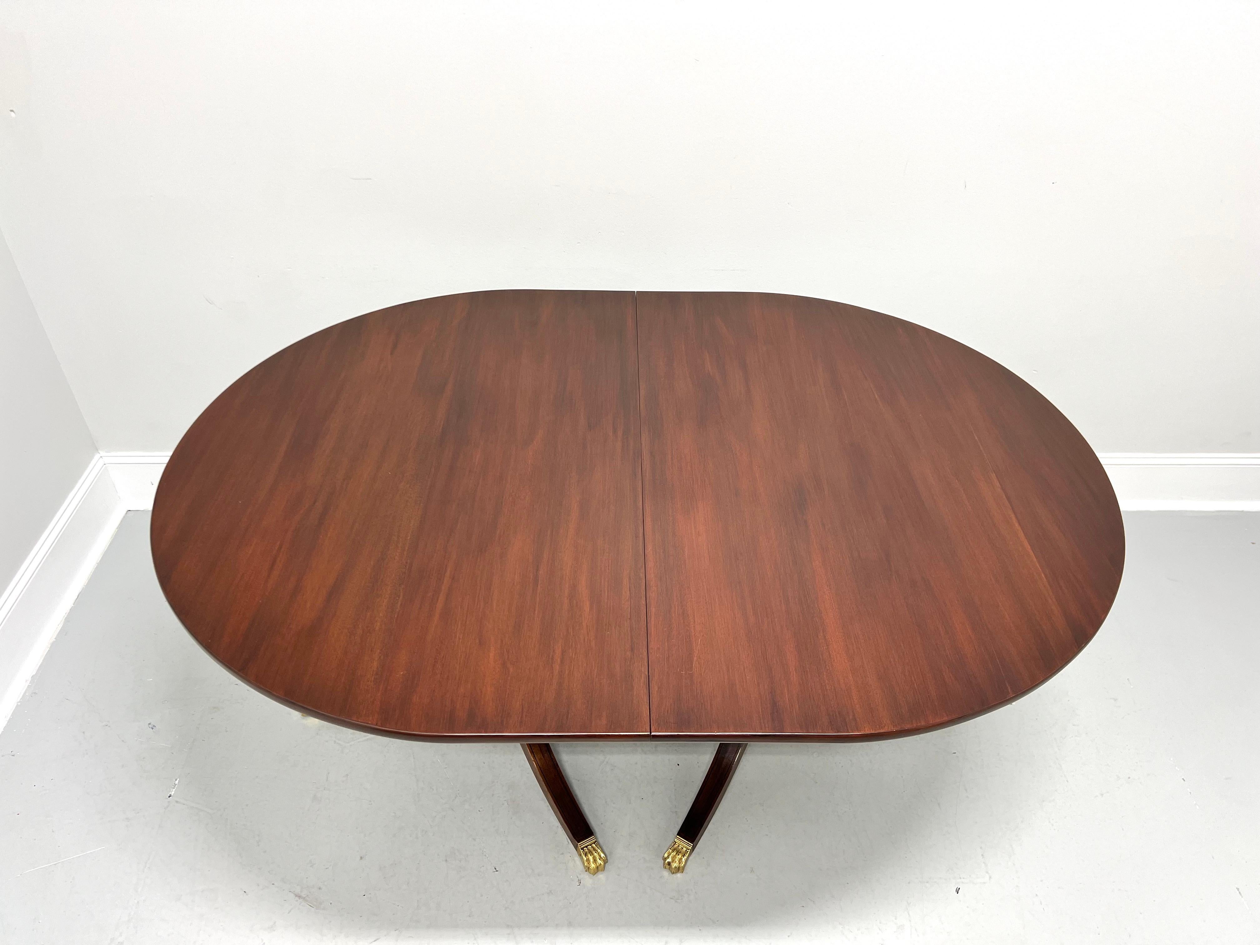 A Traditional style dining table by Henkel Harris, of Winchester, Virginia, USA. Solid mahogany, oval shaped top with a solid edge, two pedestals, each with three legs and brass paw toe caps. Includes four 12 inch extension leaves for placement on