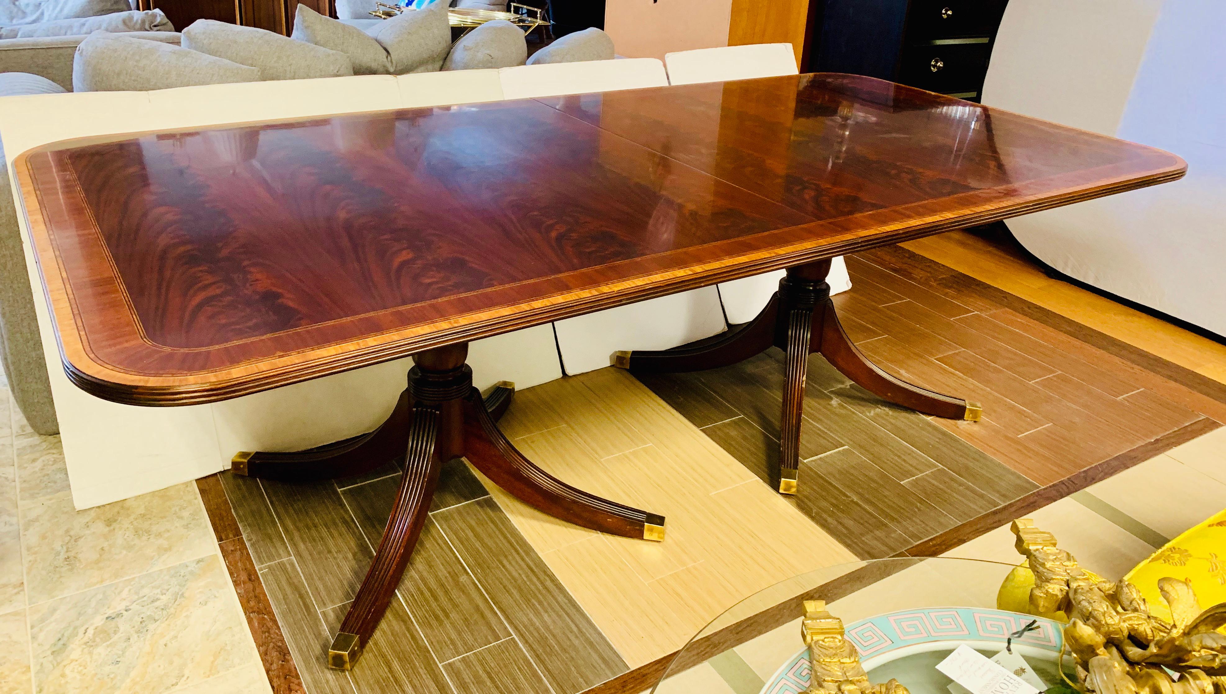 Stunning high-end book-matched flame mahogany double pedestal dining table with satinwood banded inlay. The table edge has a three beaded profile, and the pedestals have four splayed feet with solid brass casters and toe caps. Comes with two leaves
