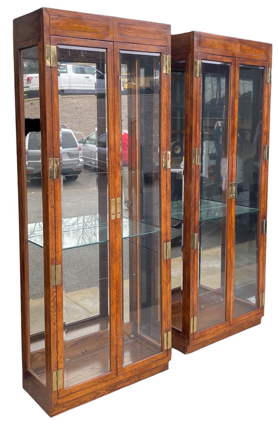 This is a pair of Henredon display cabinets with campaign styling. The backs are mirrored with adjustable glass shelves. They have two lights in each. The cabinets are marked and in very good condition. The wood is oak veneers over hardwood. 

These