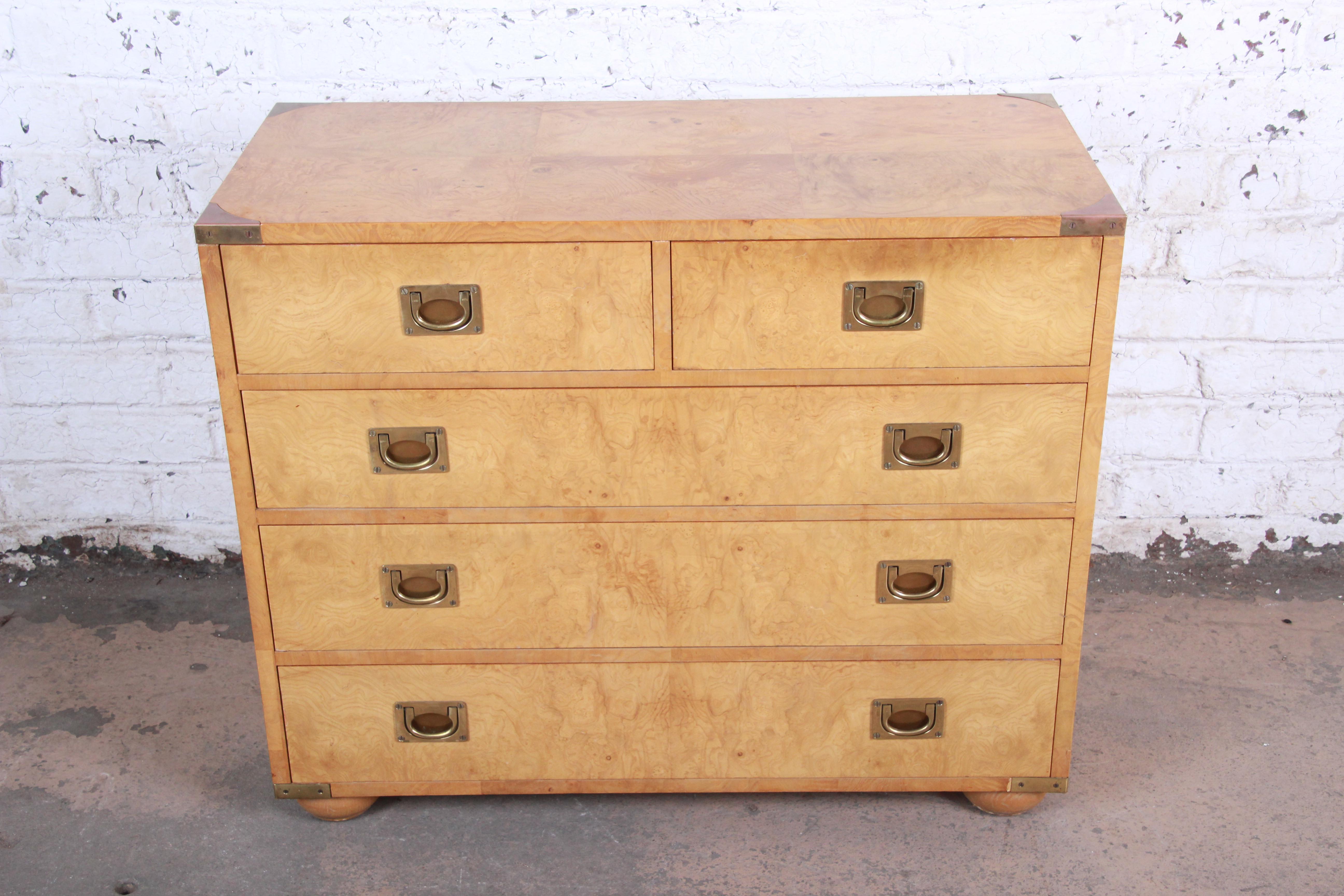 An exceptional Hollywood Regency Campaign style chest of drawers by Henredon. The dresser features stunning burl wood grain, with original brass hardware and accents. It offers ample storage, with five deep dovetailed drawers. The original label is