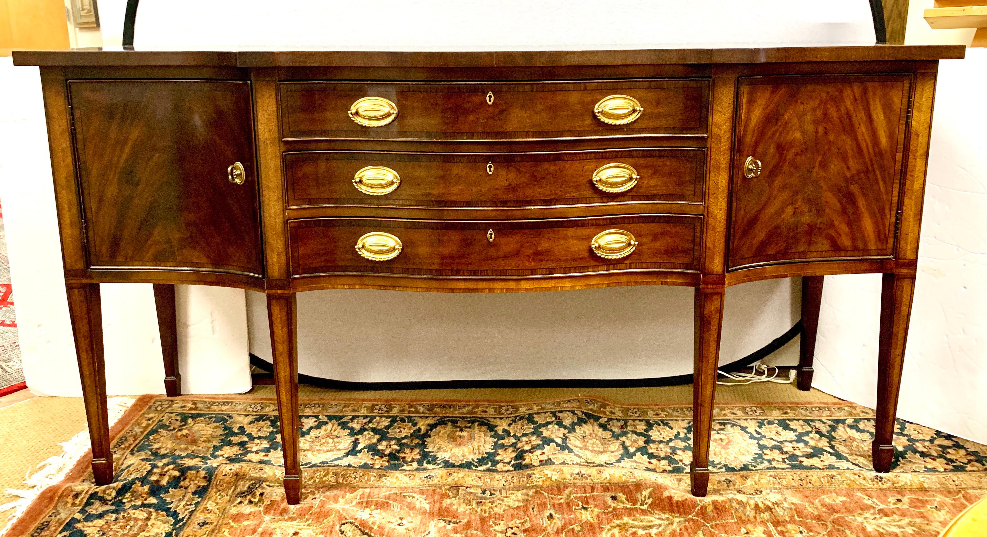 Beautifully appointed Henredon mahogany sideboard has a serpentine shaped front that is a Classic design feature and is highlighted on the top with lightly contrasting banding and bright brass hardware. There are three high quality dovetailed