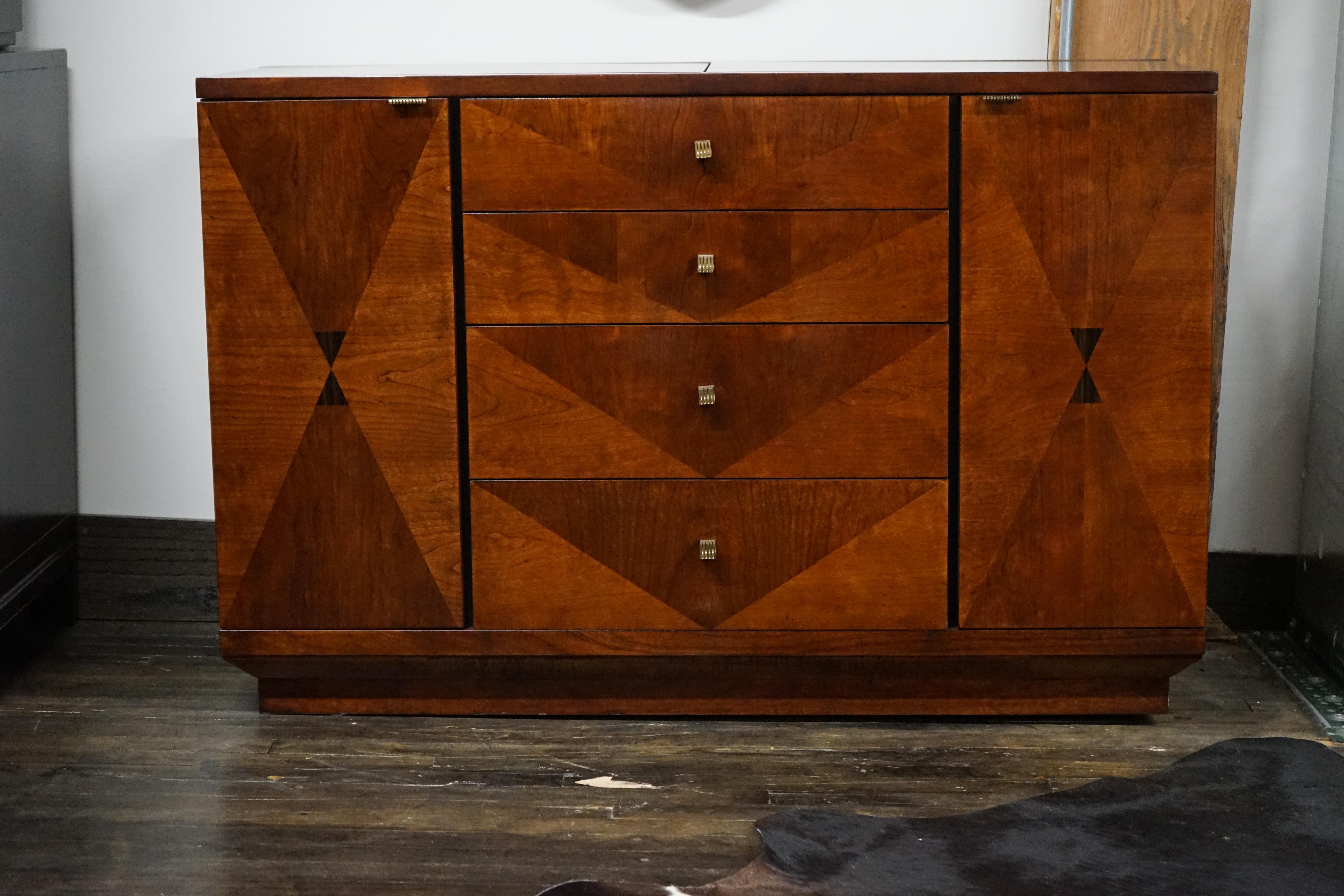 Este precioso mueble buffet/servidor/bar pertenece a la Colección Escena 4 de Hernrdon.  Presenta chapas con incrustaciones de motivos geométricos.  Los herrajes son de latón y la carcasa tiene un acabado brillante.  La parte superior se abre