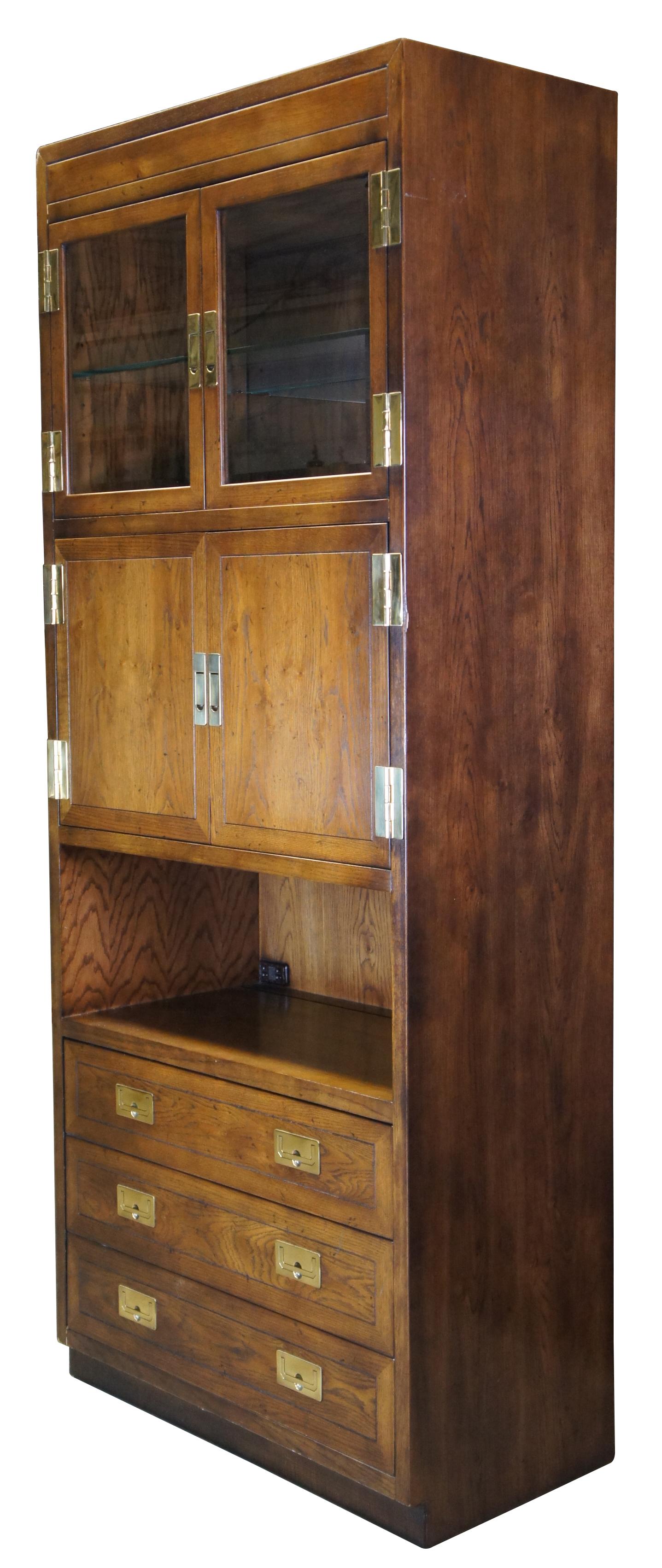Vintage Henredon campaign style cabinet. Made of oak featuring upper curio cabinet, central bar entertainment area with pull out tray and bookcase, and three lower storage drawers. All accented with modern brass hardware. Illuminated. Circa 1980s.
 