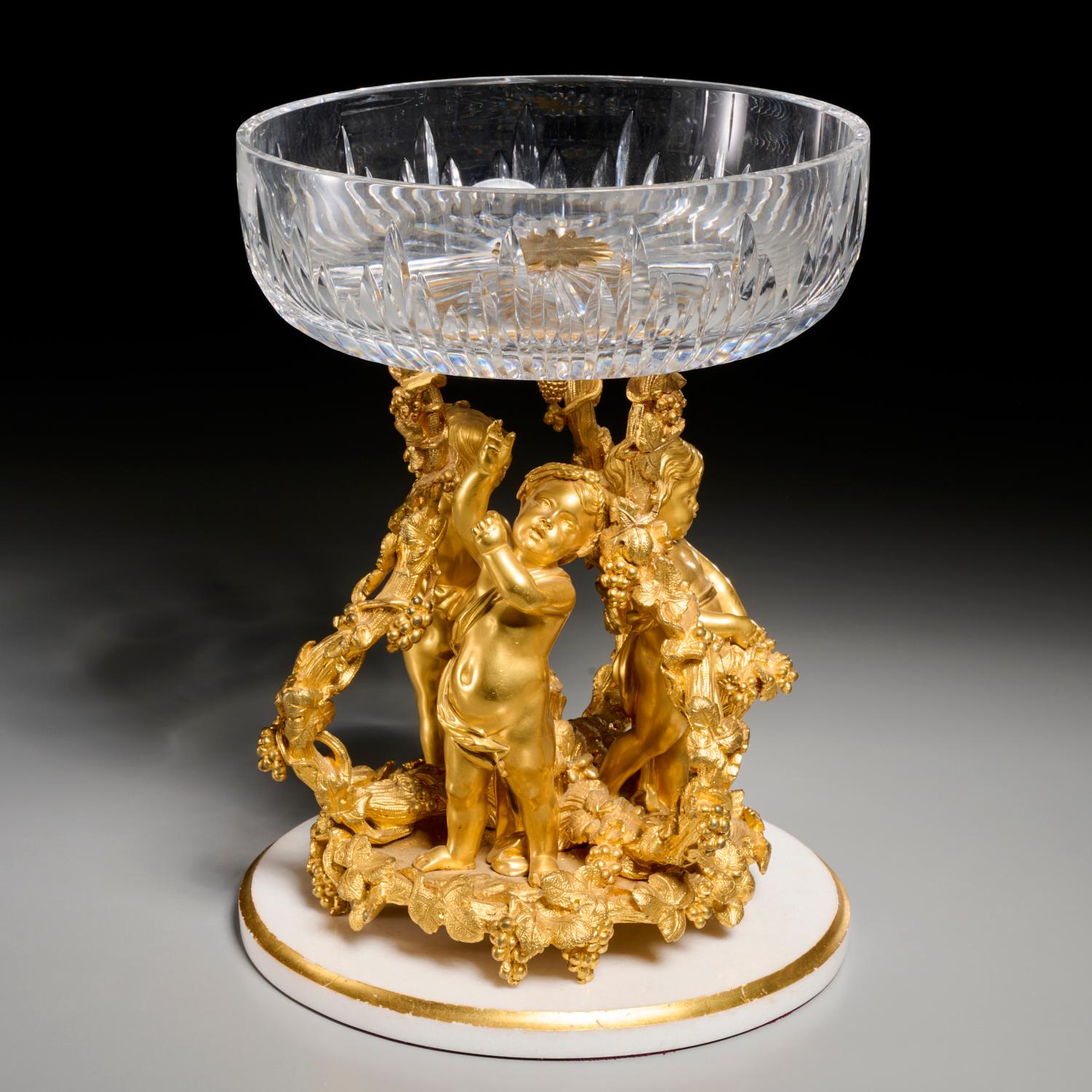After Henri Picard (French, 1831-1864), a bronze and crystal compote. The base cast has three Putti standing between fruit laden tree branches on a  white stone base with gold banding, The base is surmounted by a cut glass bowl. Henri Picard was at