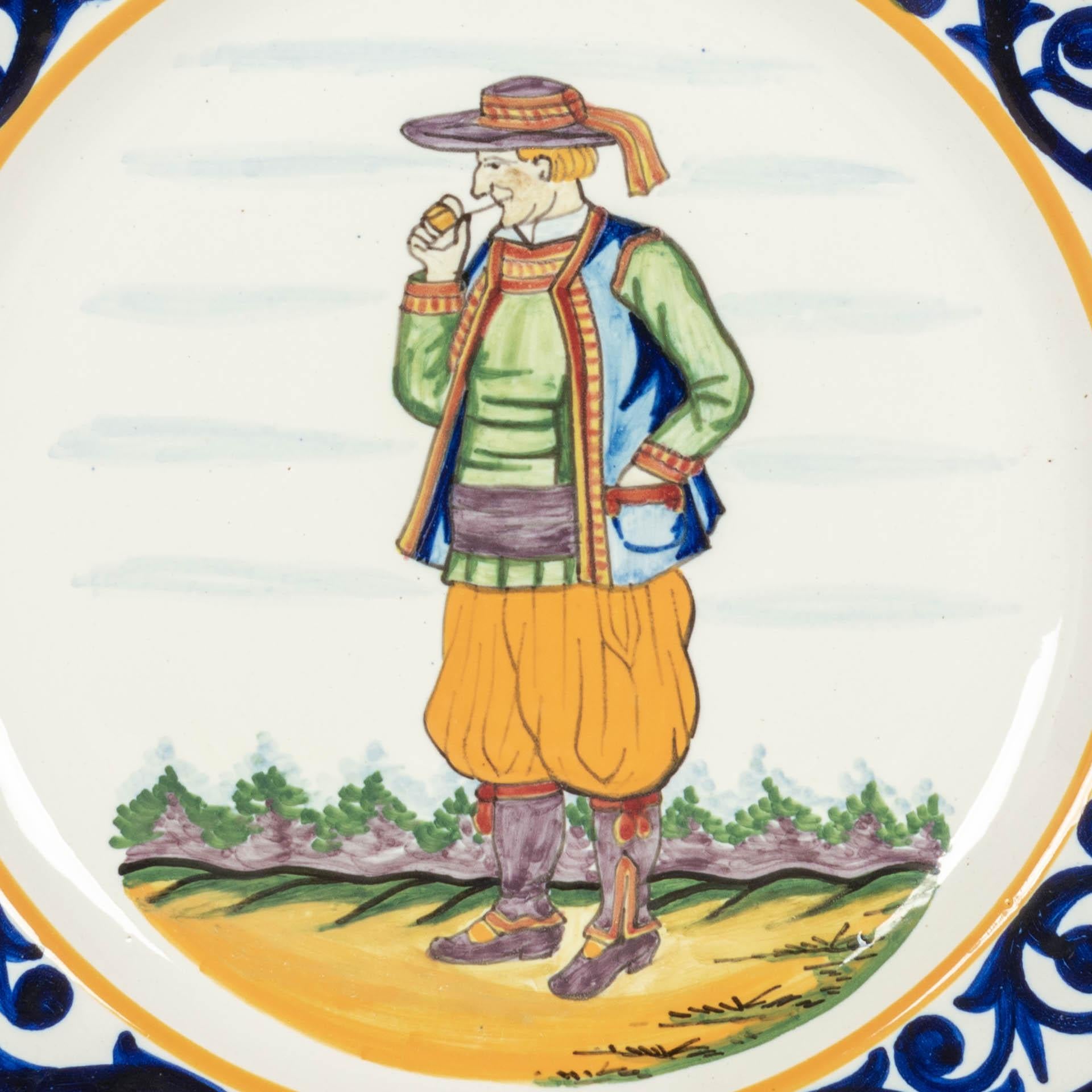 A Henriot Quimper French faience hand painted decorative plate, depicting a Breton Man with pipe in traditional clothing. Blue foliate border and with Quimper sheep crest at the top. A good quality, thick plate with minor loss to glaze. Mark on