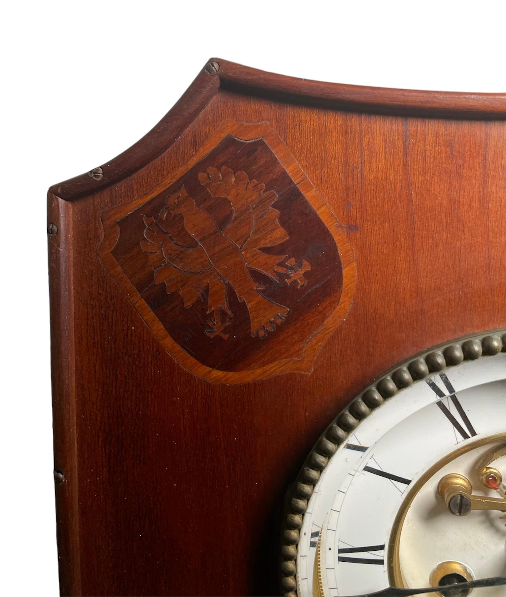 This is a Henry Lepaute wood wall clock. It depicts the clock wood case shaped as a coat of arm shield. The wood case is adorned in each one of the upper corners with one heraldic symbol made of marquetry. The round clock face is white color with