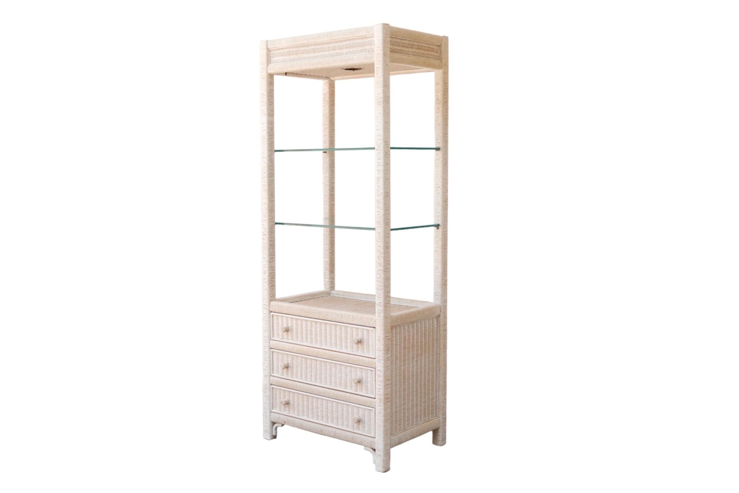 A rattan and glass cabinet designed by Henry Link for Lexington Furniture Co. Lit with a single light above two glass shelves, that rest in a white-washed rattan wrapped open frame. At the base are three drawers that open with woven rattan knob
