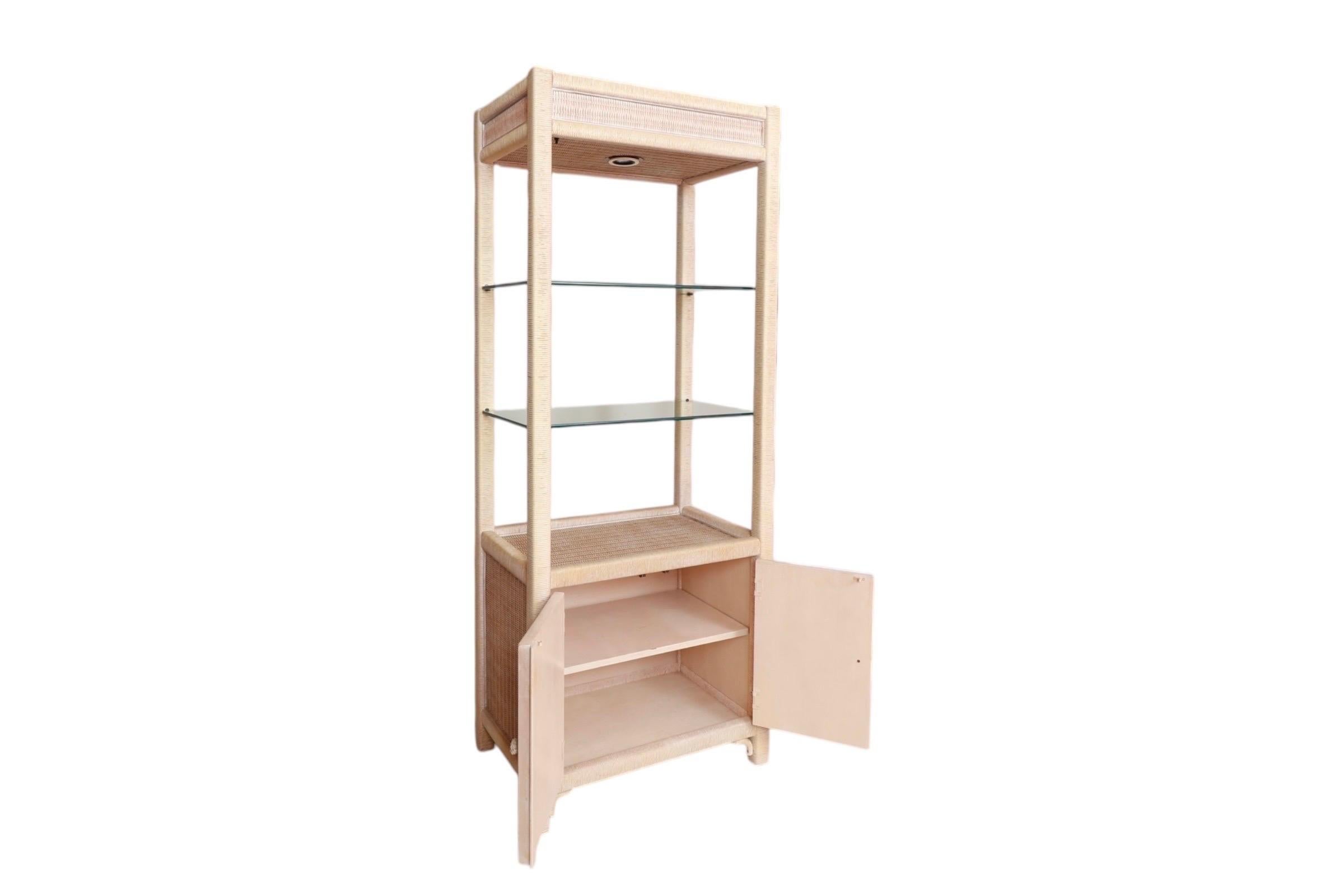 A rattan and glass cabinet designed by Henry Link for Lexington Furniture Company. Lit with a single light above two glass shelves, that rest in a white-washed rattan wrapped open frame. At the base is a cabinet that houses a single shelf, cabinet