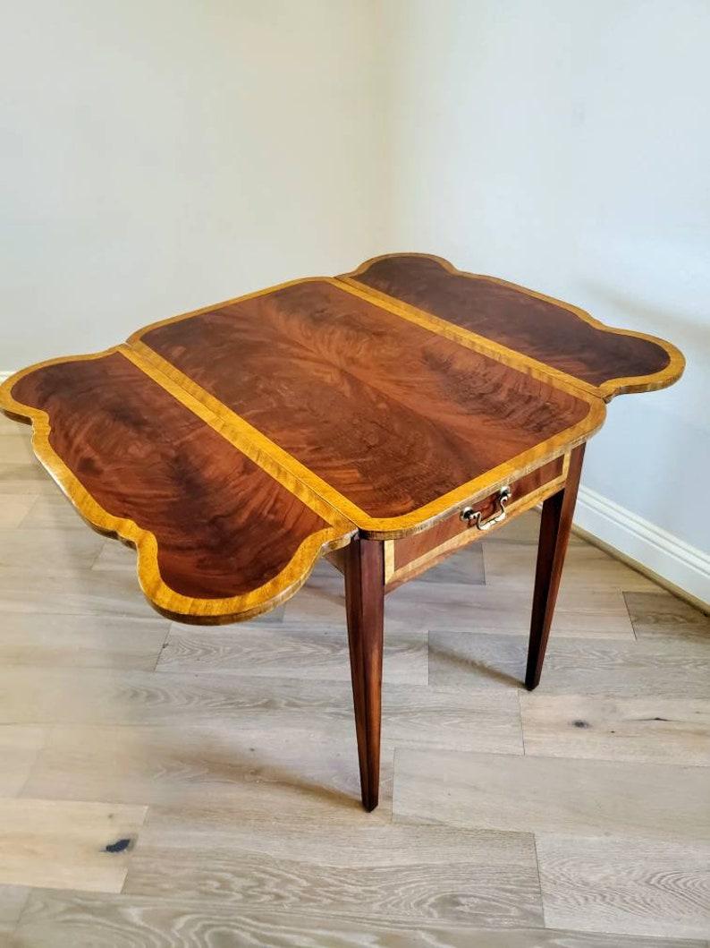 A charming Hepplewhite style mahogany pembroke table from the first half of the 20th century, having a rectangular top with serpentine shaped wooden hinged side flap extensions, outlined in a contrasting cross-banding inlay framing beautiful highly