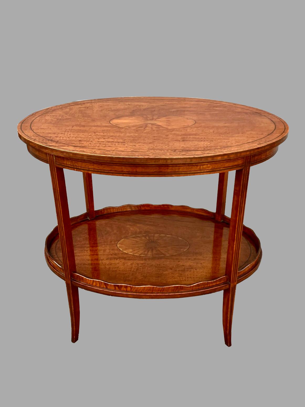 An English Hepplewhite style double tier occasional table, consisting of 2 satinwood oval surfaces each with a central boxwood inlaid paterae, the lower tier with a scalloped edge. The tops are joined with an inlaid Hepplewhite style curved leg. An