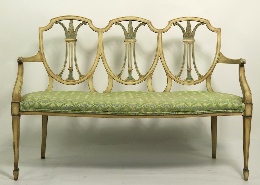 Stylish Hepplewhite style settee in decorative paint finish. The settee nhas three shied back elements, carved and curved arms, and spade feet. It is in good, original condition, shows only expected cosmetic wear to finish, normal and consistent
