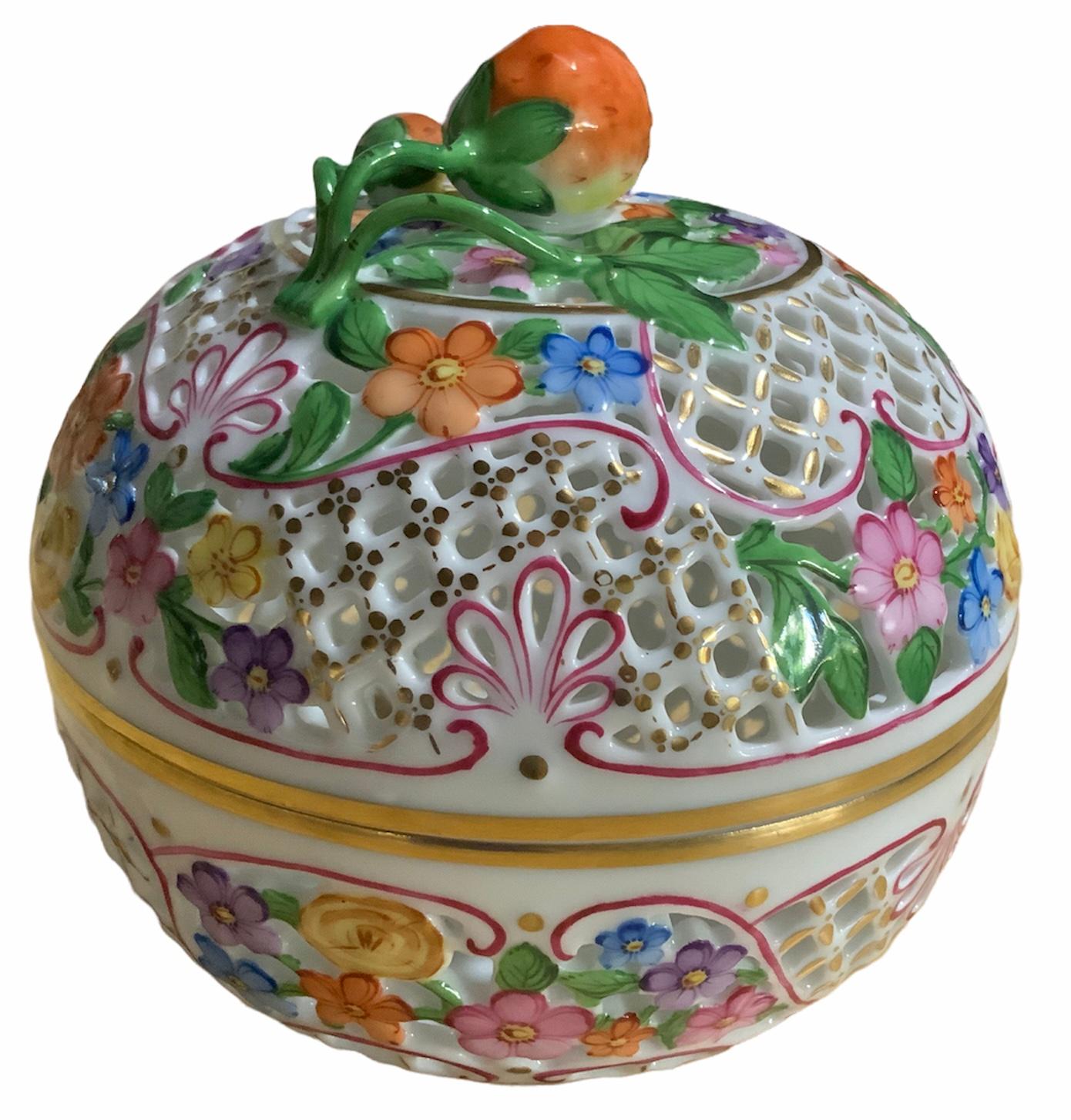 This is a Herend Porcelain reticulated round lidded potpourri bombonniere box. It is hand painted with colorful flowers and pink ribbons scrolls. Its lid and bowl borders are gilded. The lid is also decorated with a branch of two strawberries as a