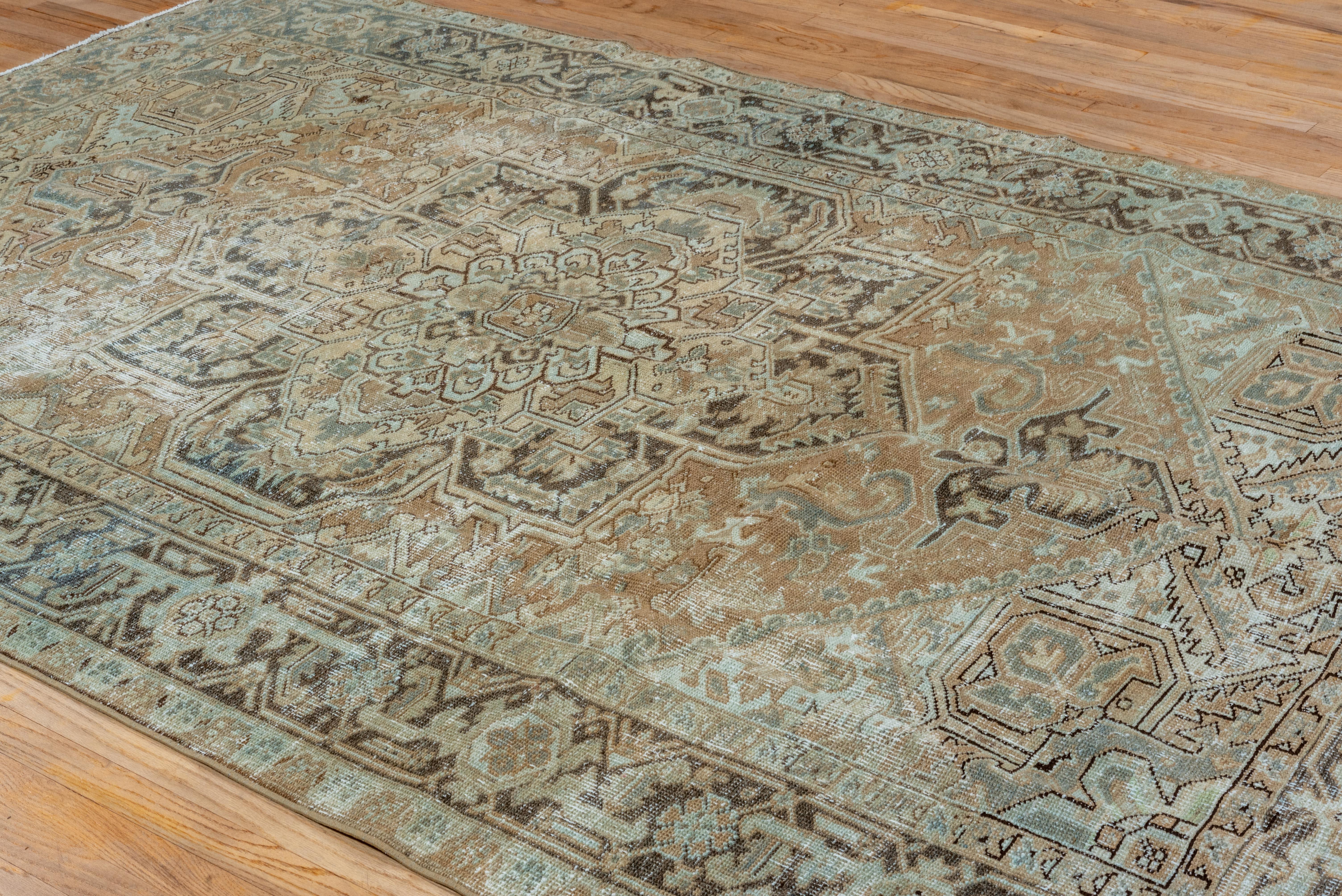This vintage Heriz rug displays a refined interpretation of the region’s bold geometric tradition, rendered in a softened palette of pale aqua, camel, sand, and muted charcoal. The central medallion anchors the composition with angular floral