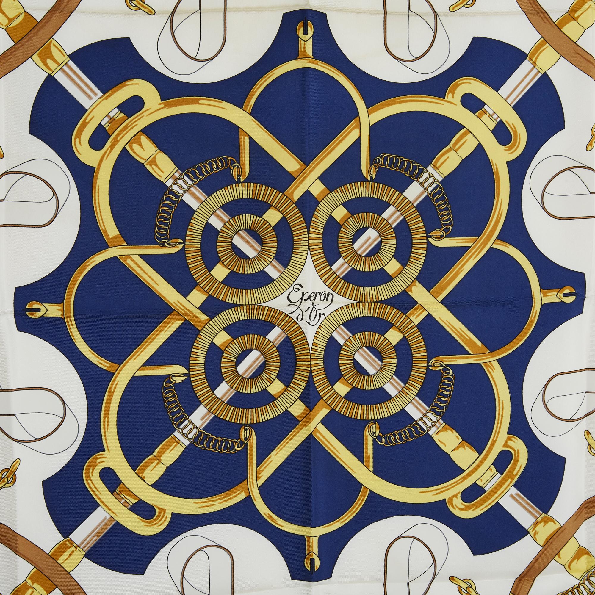 Pañuelo cuadrado Hermès 90 de sarga de seda, motivo Eperon d'or de Henri D'Origny, publicado en 1974 y reeditado en 1989 (éste), motivos en tonos crudos y dorados, bordes azul marino. Ancho 90 cm x largo 90 cm aproximadamente. El pañuelo es vintage