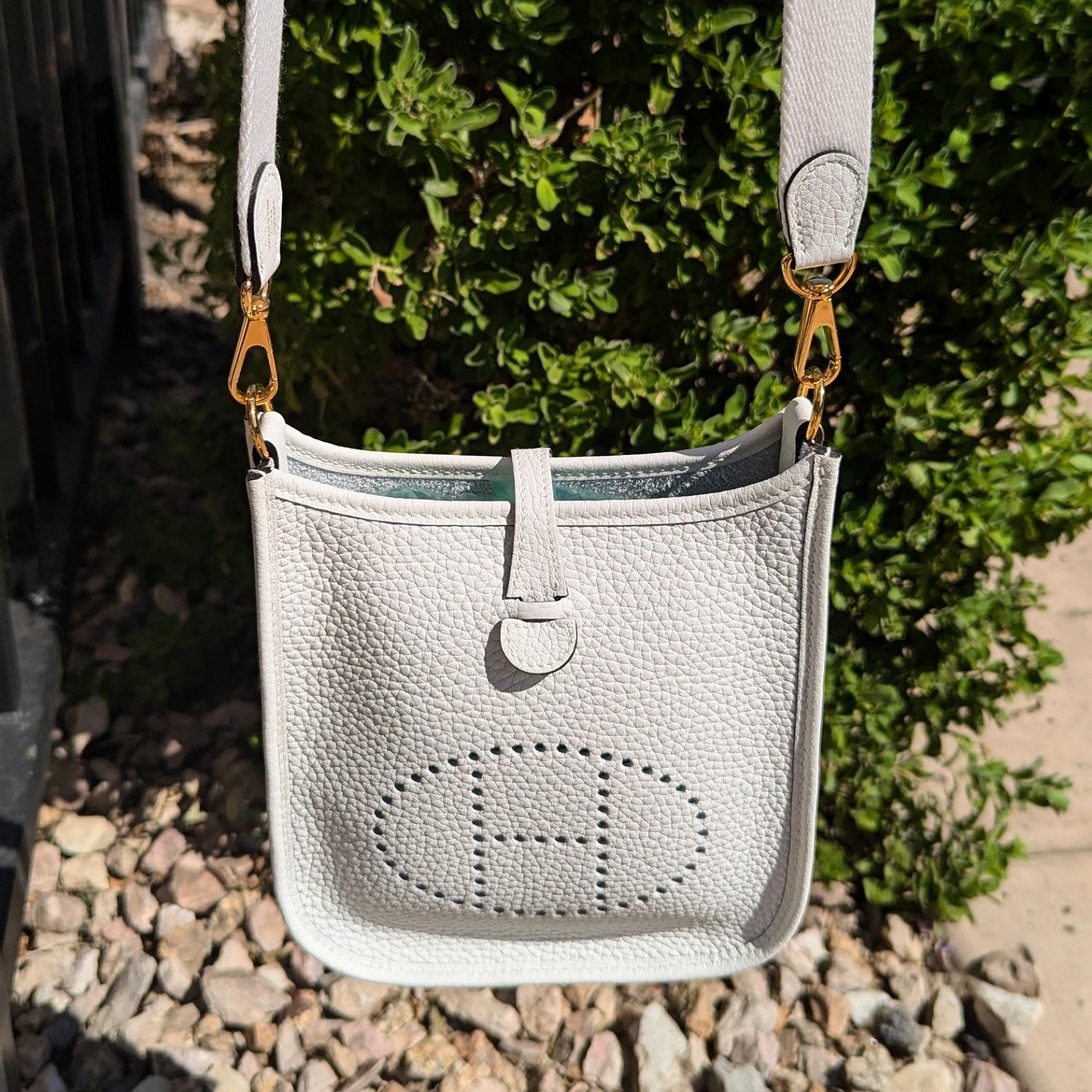 Hermès Taurillon Clemence Evelyne TPM in New White. Questa borsa messenger nella misura più piccola è realizzata in pelle di vitello Taurillon. La borsa presenta una tracolla in tela bianca, finiture dorate lucide e un logo ovale Hermes H. H.
