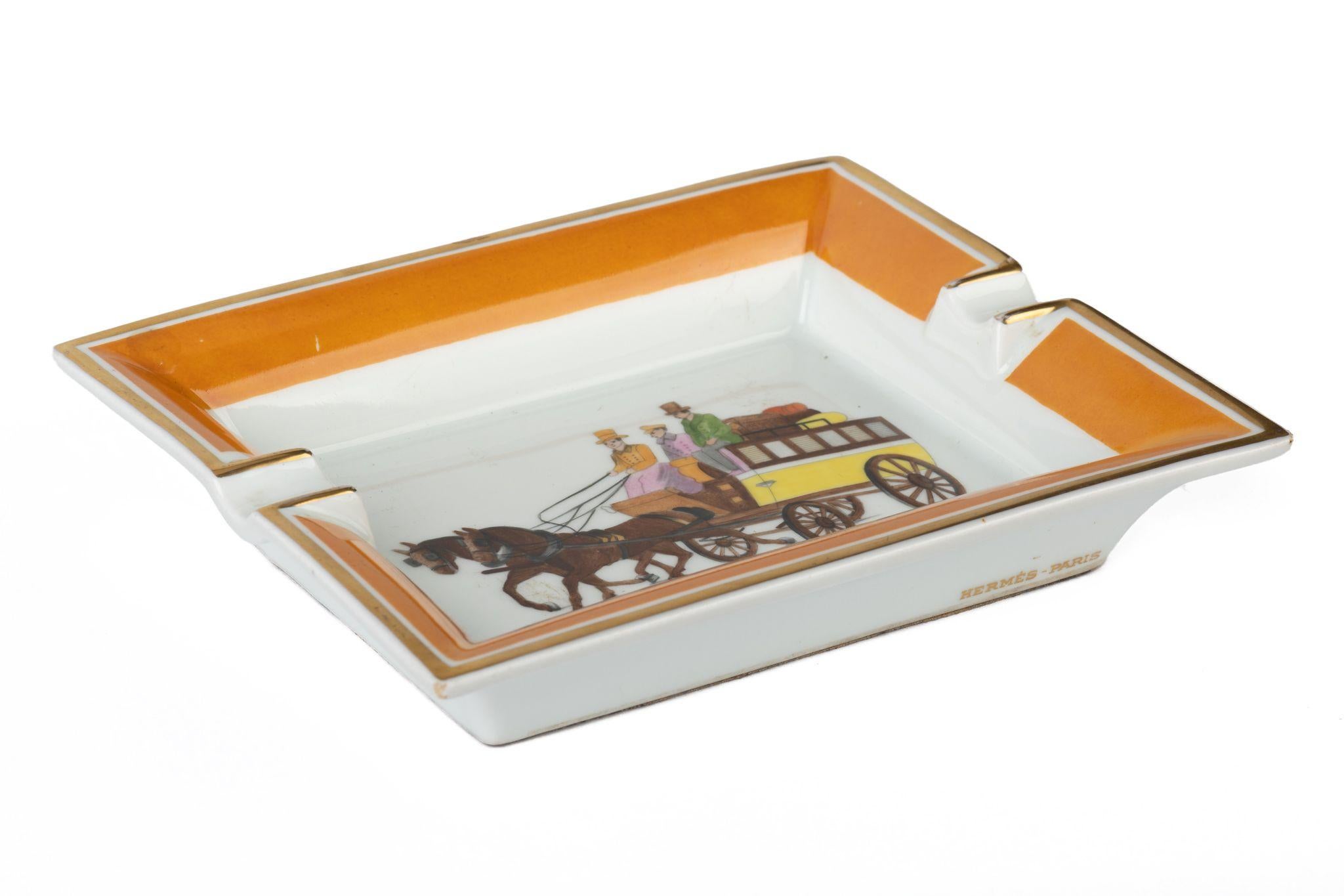 Hermès signature ashtray in white porcelain with a horse carriage design in yellow and brown. The edges are colored orange. Suede stamped bottom.
