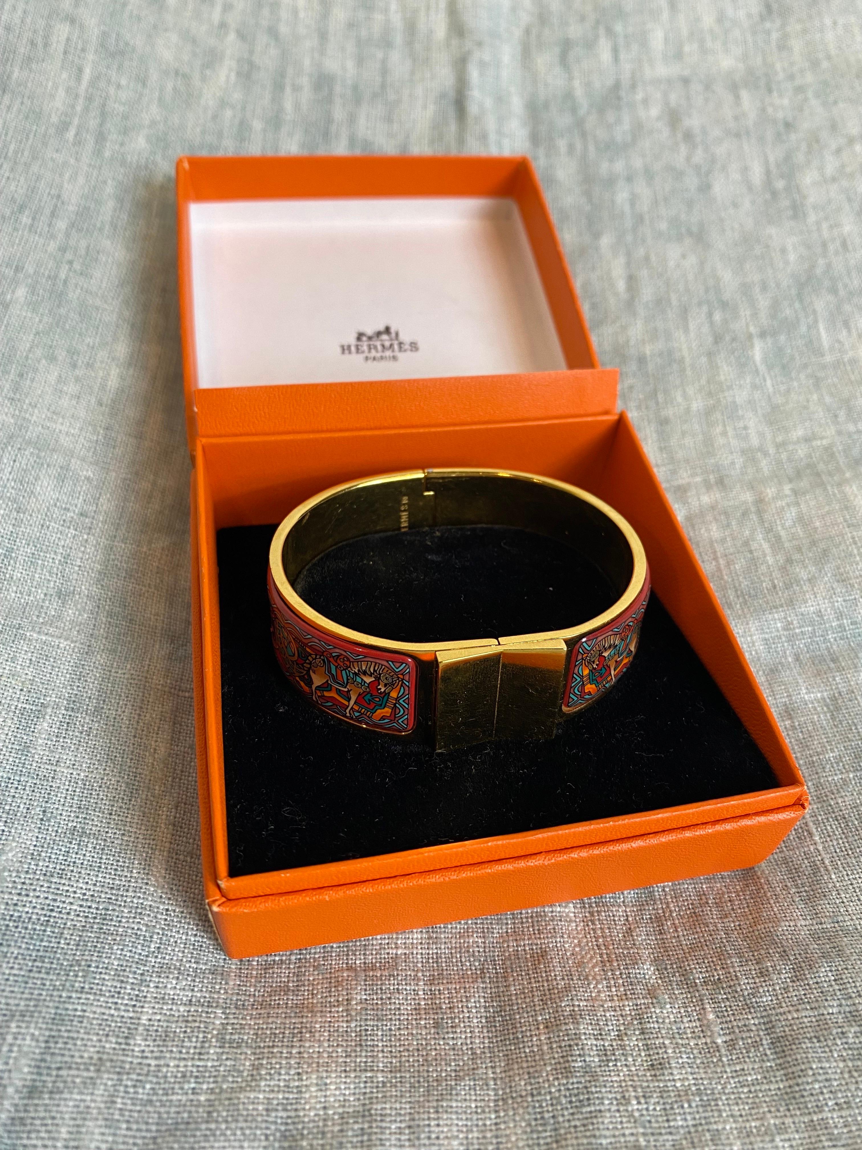 Bracciale bangle di Hermes in metallo dorato con disegni in smalto multicolore. Chiusura a strappo, girando il quadrato anteriore verso l'alto. 
In ottime condizioni, presenta alcuni graffi di utilizzo nella parte metallica. 
Con scatola originale