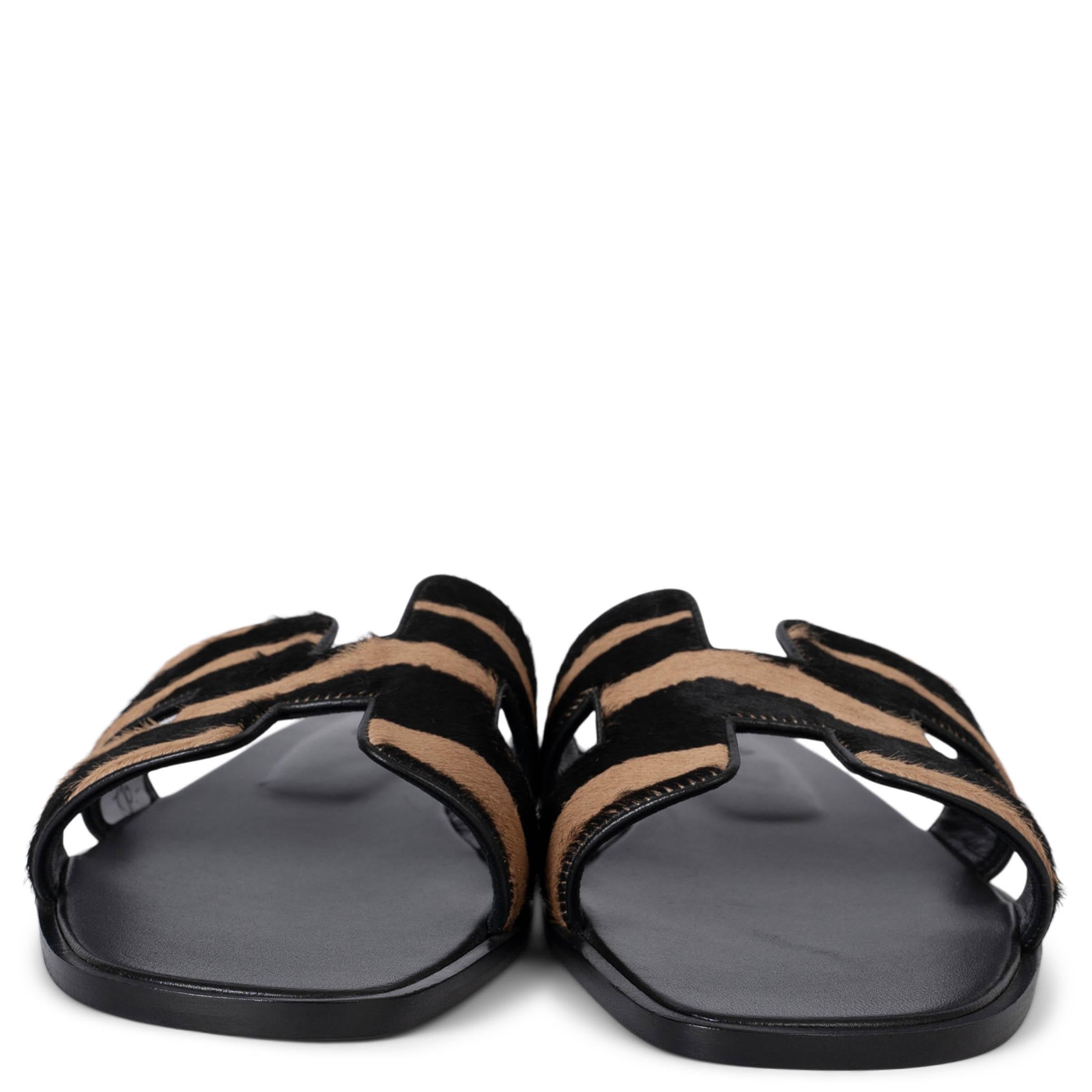 These 100% authentic Hermès Oran slide sandals are crafted from beige and black calf hair, featuring the iconic H cut-out design. They are finished with black leather soles, offering a chic and timeless look perfect for elevating any casual outfit.