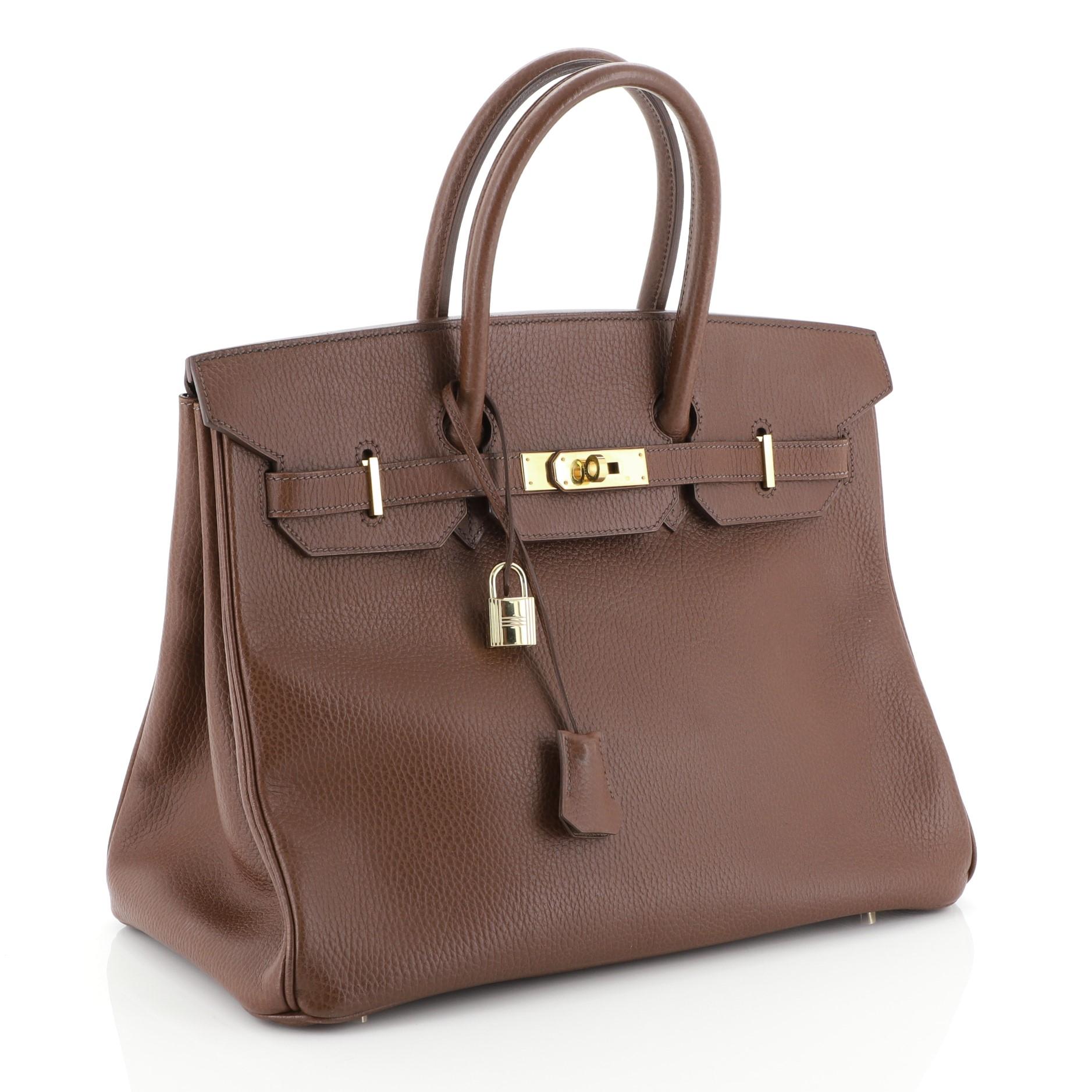 This Hermes Birkin Handbag Marron Fonce Ardennes with Gold Hardware 35, crafted in Marron Fonce brown Ardennes leather, features dual rolled handles, frontal flap, and gold hardware. Its turn-lock closure opens to a Marron Fonce brown Chevre leather