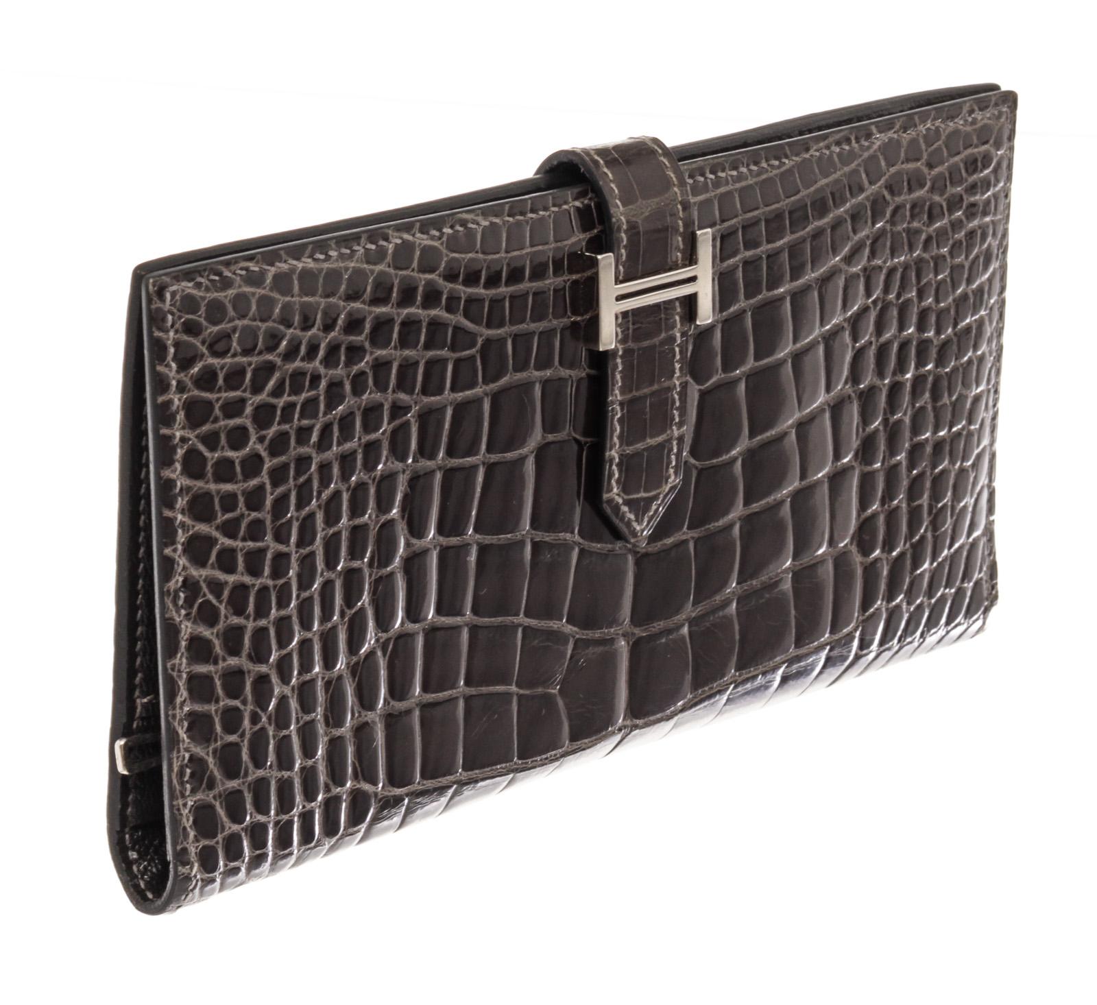 Hermes Bearn Wallet is cone in shiny black Alligator leather, featuring a bifold design and a silver-tone strap closure. The interior is crafter in black Chevre Leather, featuring five card slip pockets, two slip pockets for paper currency, one zip
