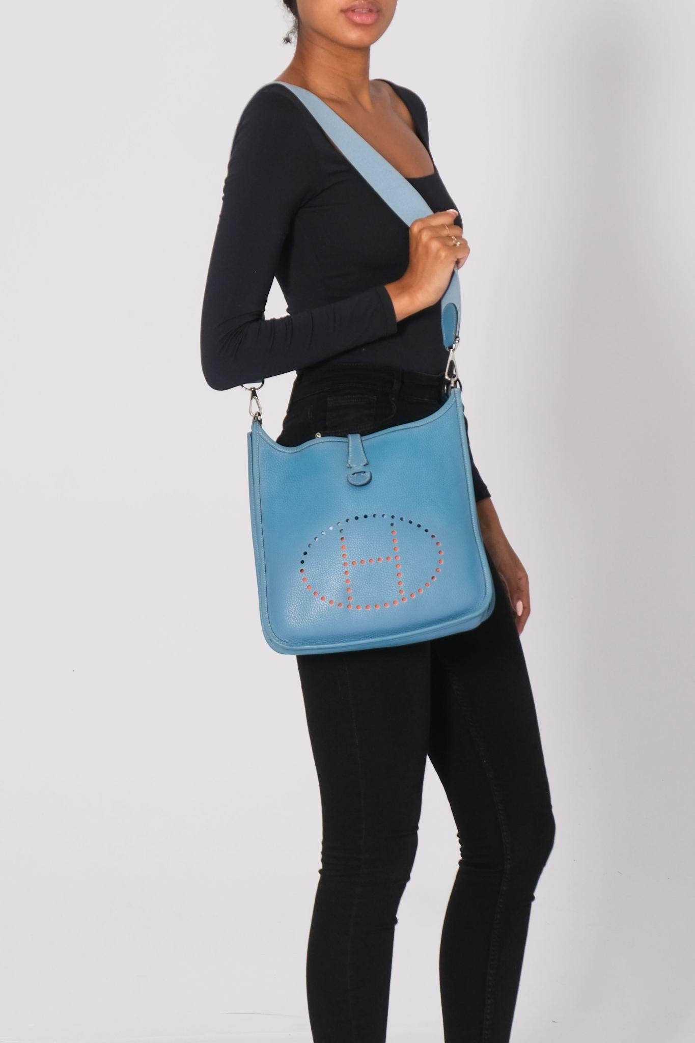 Hermes Evelyn bag size pm made of textured leather. The shoulder bag features a canvas strap, a front perforated Hermes H, and light palladium hardware including clasps for the shoulder strap. The bag has an open top and a matching suede interior.