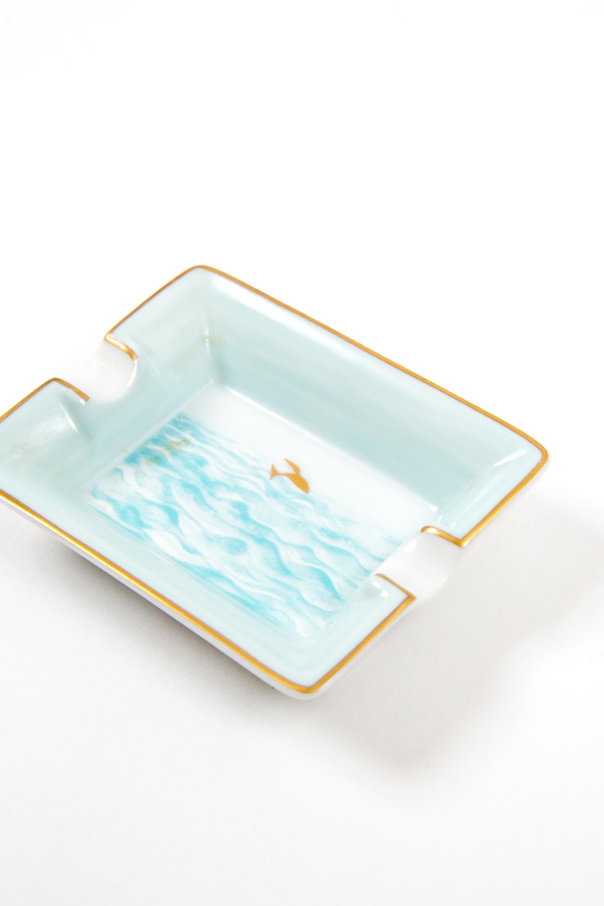 Hermès Blue Mini “Gold fish in the Ocean” porcelain de Limoges ashtray organizer featuring a fish motive, a blue and gold border, a gold brush finishing, a Hermes gold-tone side signature. 
Circa 1980s 
In good vintage condition. 
Maxi Length 2.9in