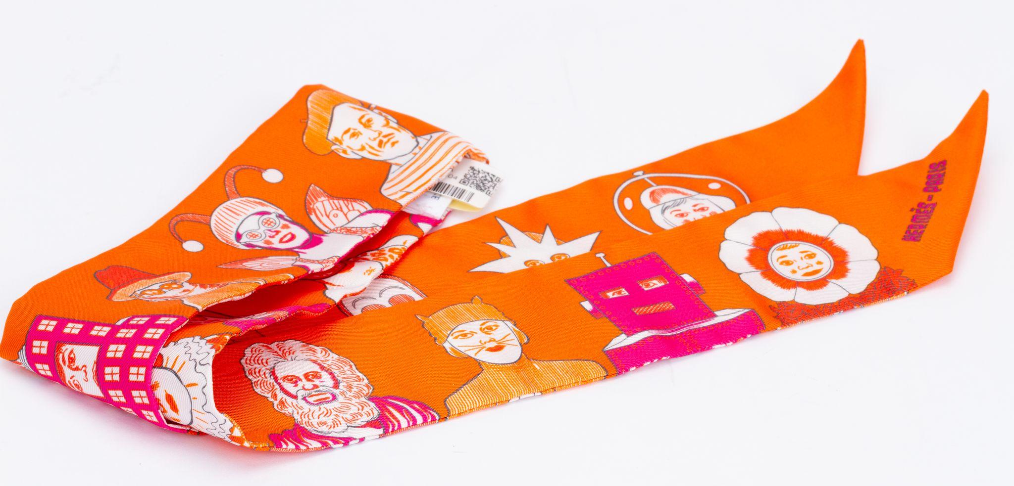 Nouvelle édition Hermès Dresscode twilly. L'écharpe est fabriquée en soie et se présente dans un orange vibrant. L'impression montre différents personnages, par exemple la statue de la liberté. La pièce est livrée dans sa boîte et sa pochette