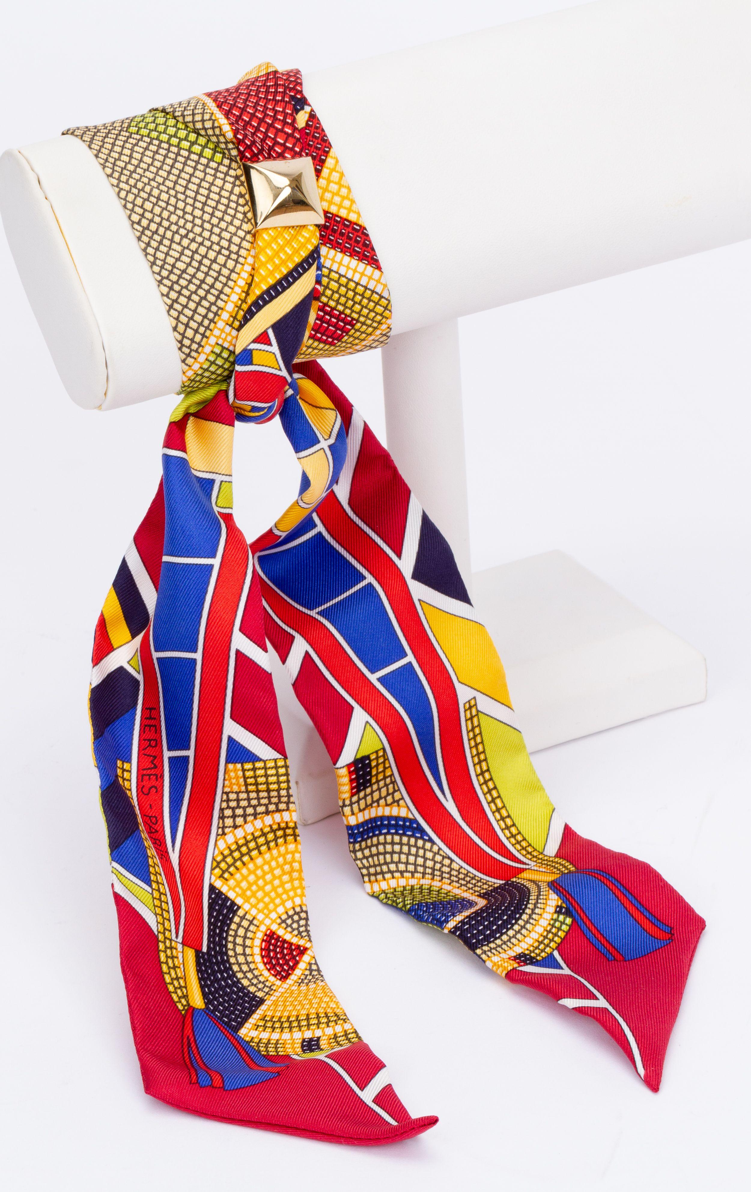 New Hermès Geometric Twilly. The scarf is made out of silk and is multicolored. The print shows different geometrical figures and forms. The pieces comes with a scarf ring, the original box and ribbon.