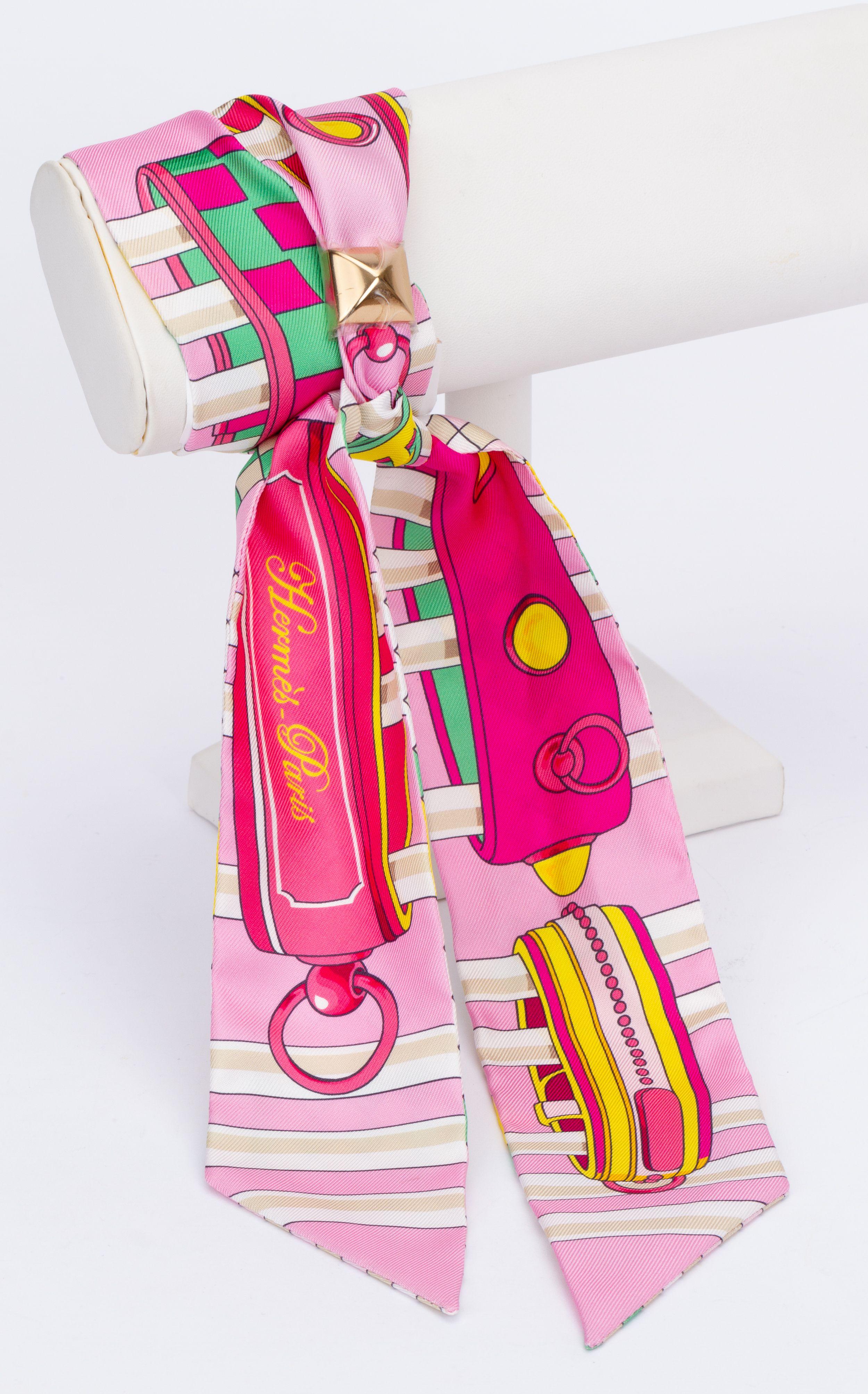 New Hermès Trophy Ribbon Twilly. The scarf is made out of silk and comes in the color pink. The print shows different geometrical forms and trophy ribbons. The pieces comes with a scarf ring and is packed in the original box.
