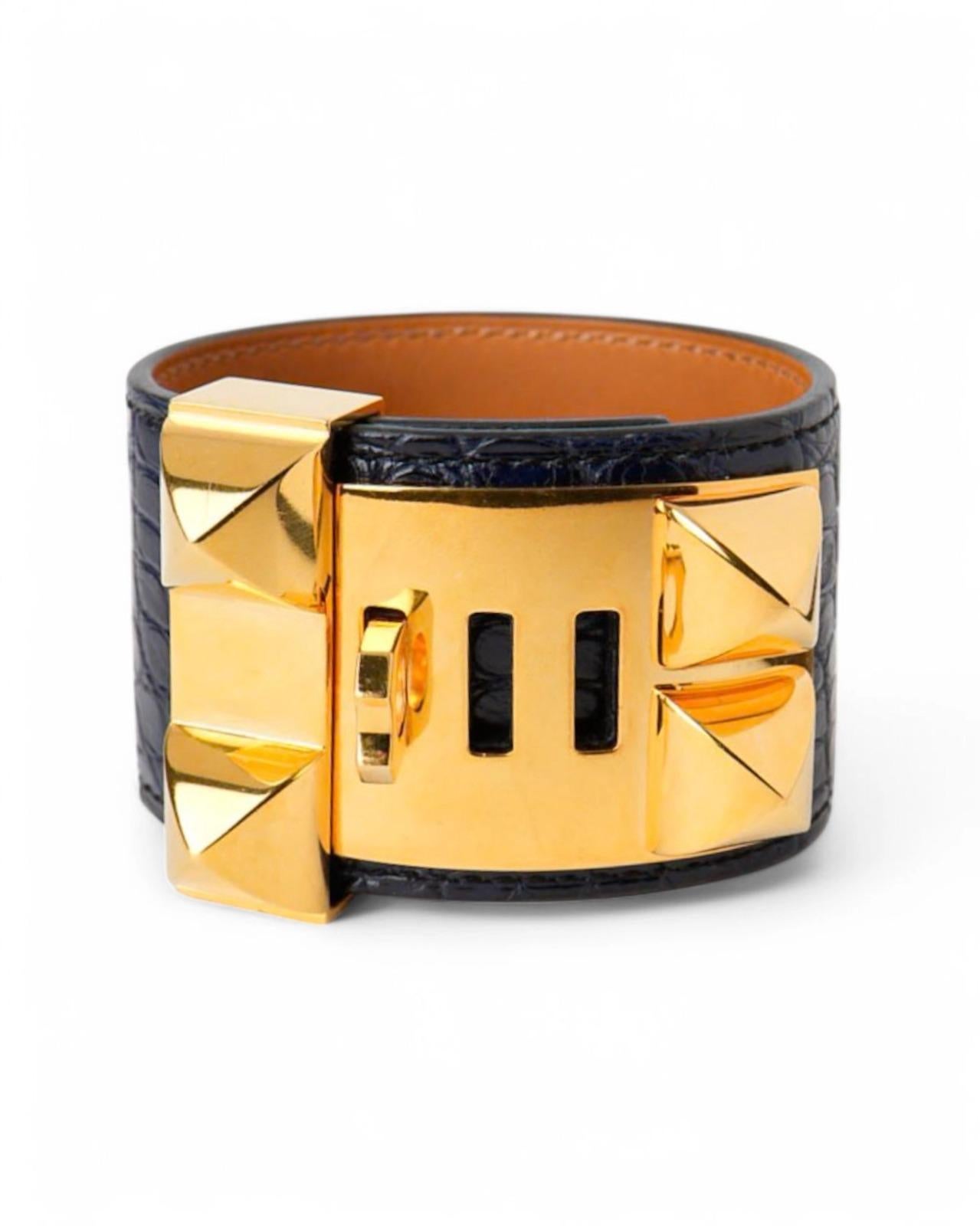 Bracciale firmato Hermès, modello Collier De Chien, realizzato in pregiata pelle di alligatore blu e arricchito da borchie dorate. Questo bracciale regolabile offre un design audace e sofisticato, perfetto per aggiungere un tocco di lusso a