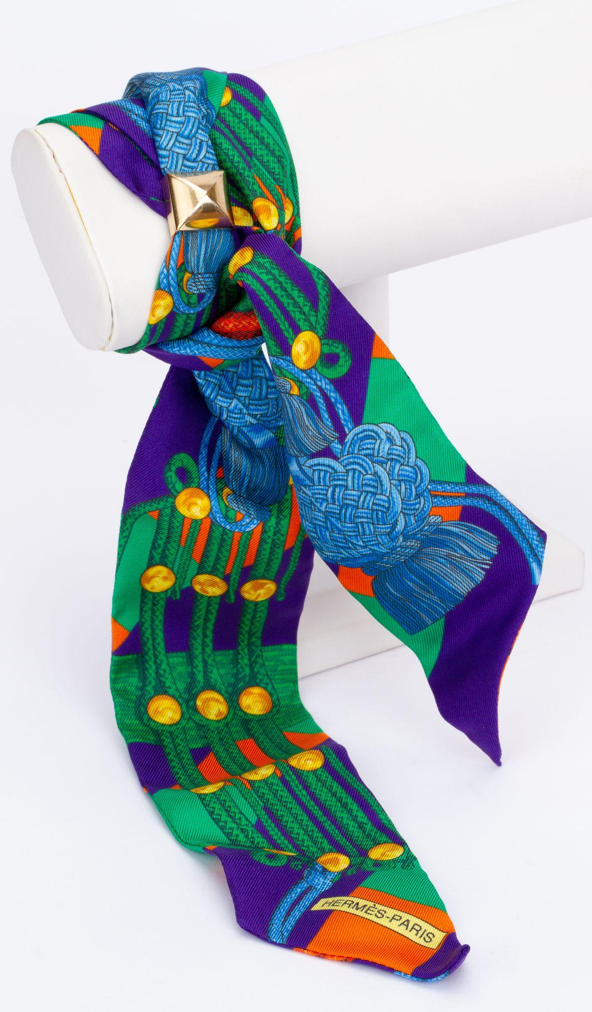 New Hermès Brandebourg Twilly. The scarf is made out of silk and the main color is a green/blue. The print shows the buttons of a military jacket. The piece comes with a scarf ring. It is packed in a original box and ribbon.
