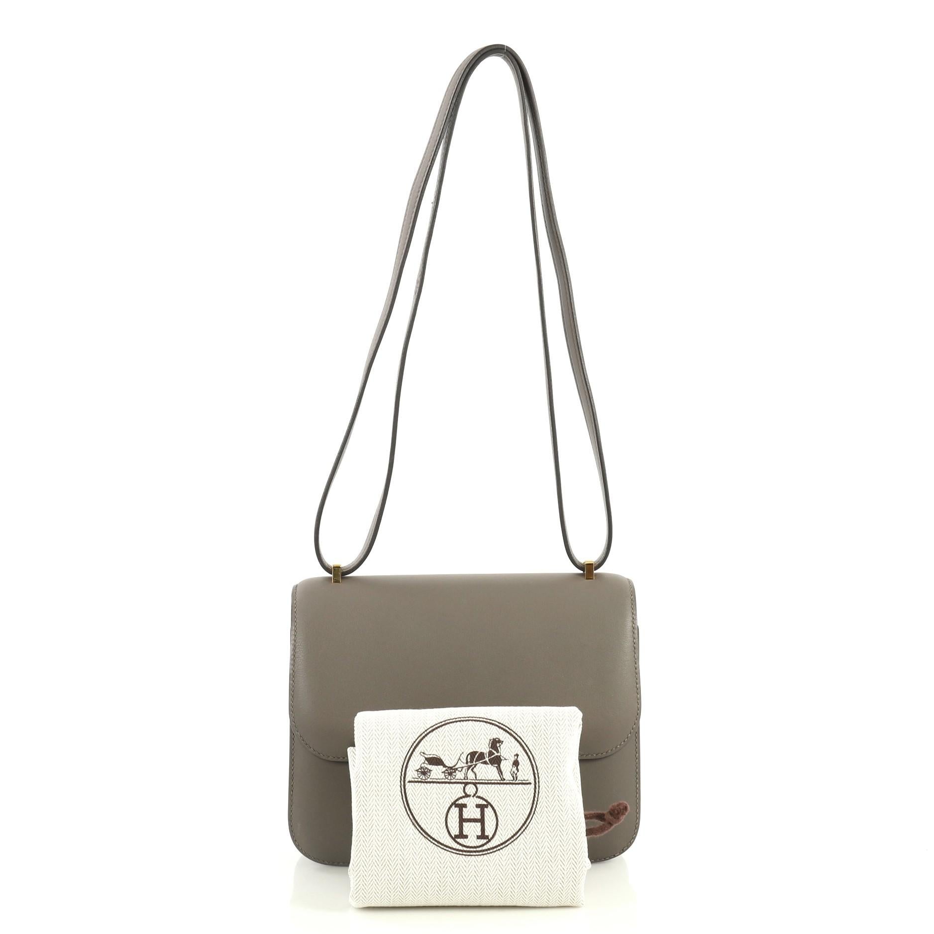 This Hermes Constance Handbag Swift 18, crafted from Etain gray Swift leather, features a long leather strap and gold hardware. Its push-lock closure opens to an Etain gray Swift leather interior with two open compartments and side slip pockets.