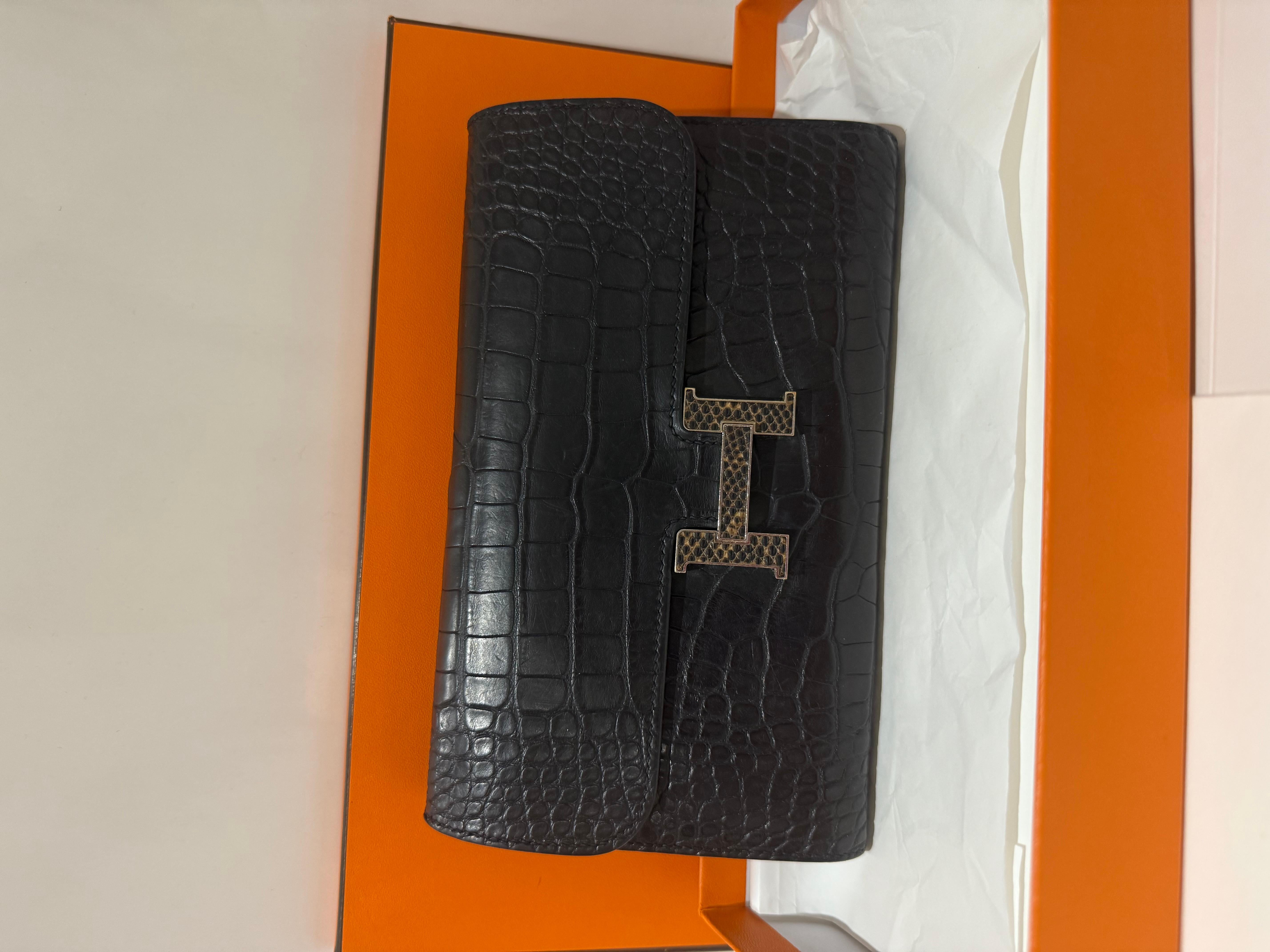Portefeuille long Hermès Constance en cuir d'alligator noir mat avec boucle en lézard Ombre. Ce portefeuille est finement confectionné en cuir d'alligator mat de couleur noire. Le rabat avant s'ouvre avec un 