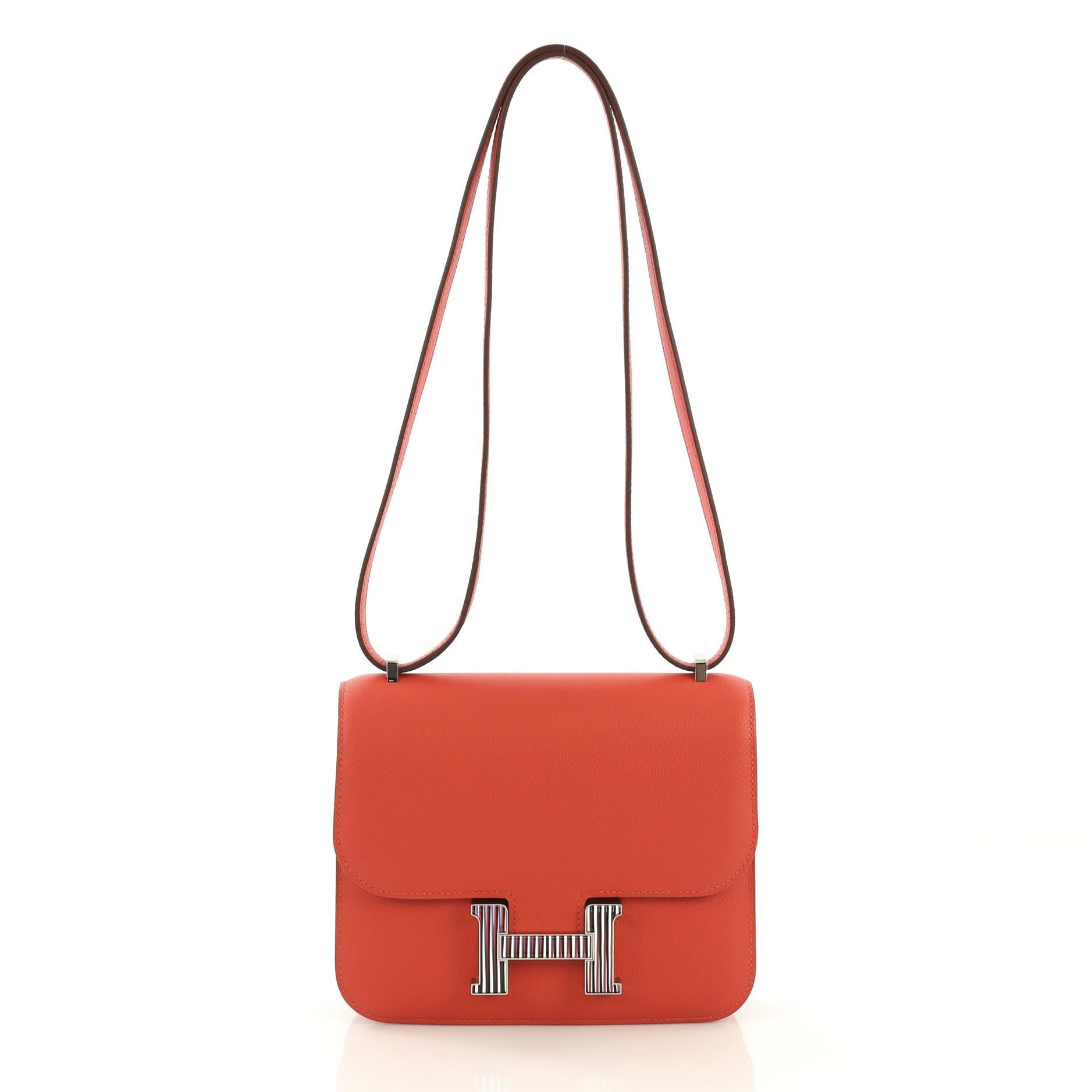 This Hermes Constance Optique Handbag Evercolor 18, crafted from Capucine red Evercolor leather, features flat leather strap, frontal flap, and enamel optique hardware. Its push-lock closure opens to a Capucine red Agneau leather interior with two