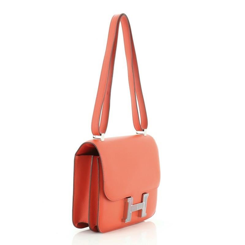 This Hermes Constance Optique Handbag Evercolor 24, crafted from Capucine red Evercolor leather, features flat leather strap, frontal flap, and enamel and palladium hardware. Its push-lock closure opens to a Capucine red Agneau leather interior with