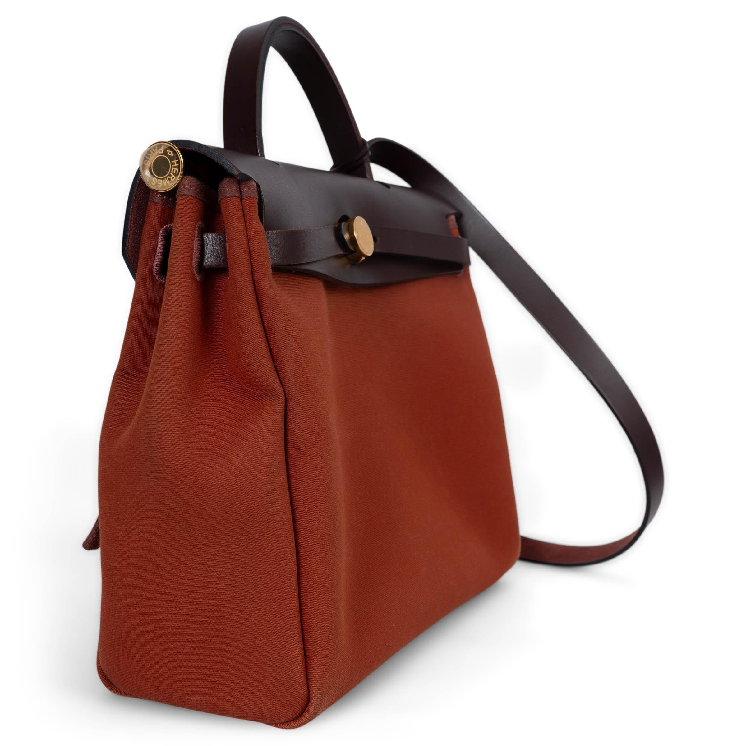 This 100% authentic Hermès Herbag Zip 31 Retourné Verso bag is crafted from Rouge Sellier (Maroon) Vache Hunter leather and Cuivre (dark brick) Toile Officier, accented with a vibrant Orange Mécano (coral) zip pocket on the back. It features elegant