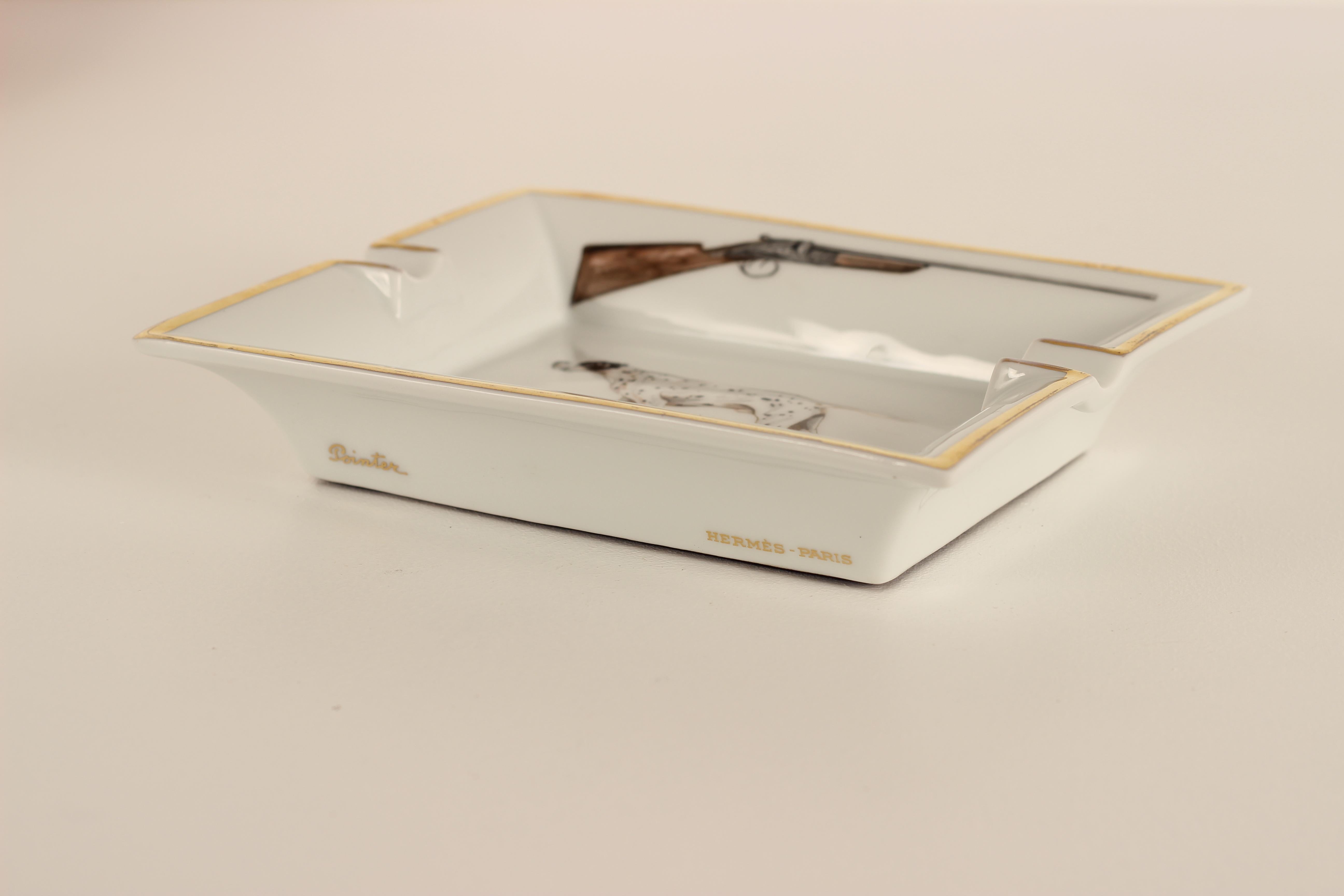 A Porcelain ashtray or vide Poche Catchall by renowned Fashion House Hermes. 

Featuring two hunting guns and a Pointer Dog mid pose. Surrounded by a gilt border and signed on one of the faces of the ashtray, this piece has no chips or cracks, but