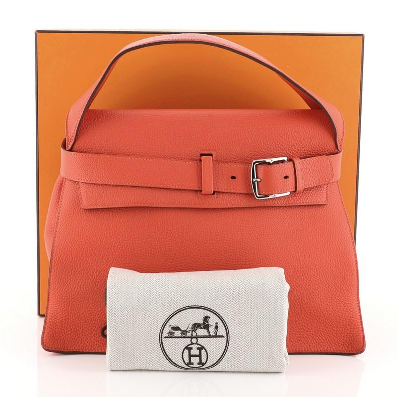 This Hermes Etribelt Handbag Togo, crafted in Capucine red Togo leather, features a wide leather strap, front belt detail and palladium hardware. Its buckle closure opens to a Capucine red Chevre leather interior with side slip pocket. Date stamp