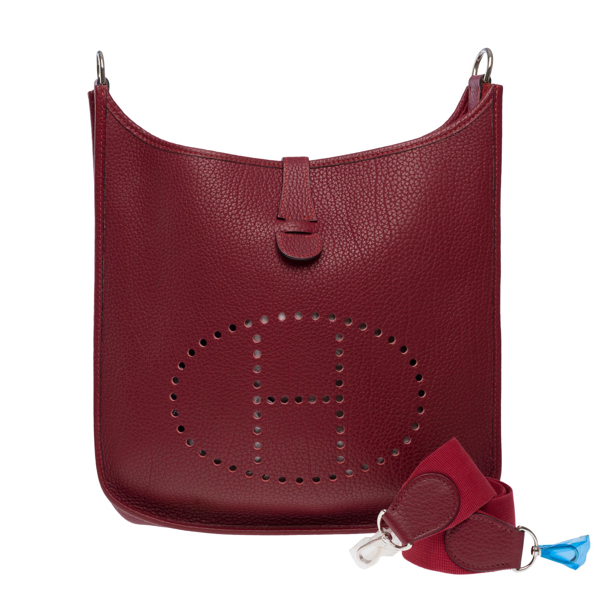 Sac à bandoulière iconique Hermès Evelyne GM en cuir Fjord Rouge H (bordeaux), quincaillerie en palladium argenté, bandoulière amovible en toile rouge pour un portage à l'épaule ou en travers du corps.

Fermeture par bouton-pression sur le