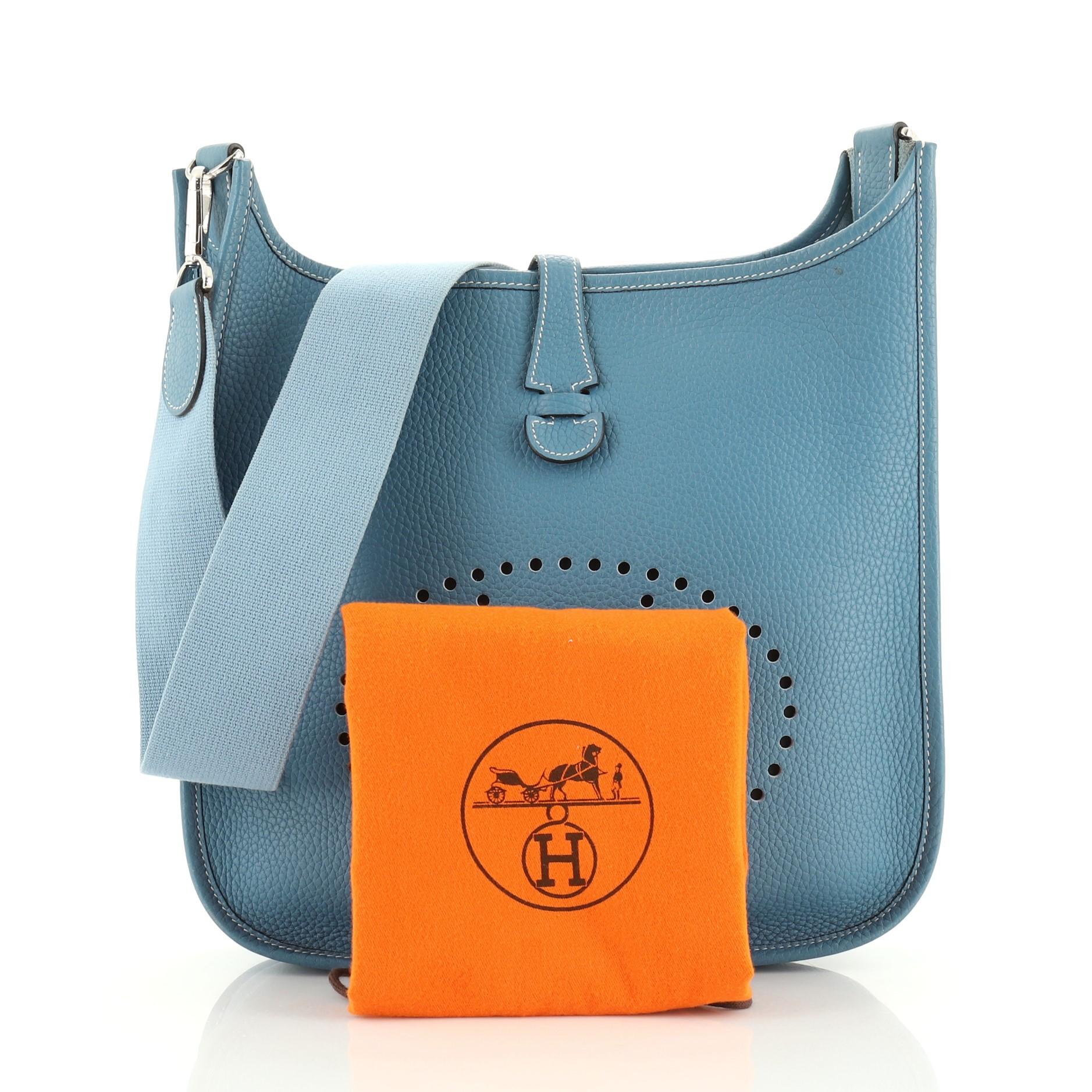 This Hermes Evelyne Crossbody Gen I Clemence PM, crafted in Bleu Jean blue Clemence leather, features a textile shoulder strap, perforated H design at the front, and palladium hardware. Its top leather tab closure opens to a blue raw leather