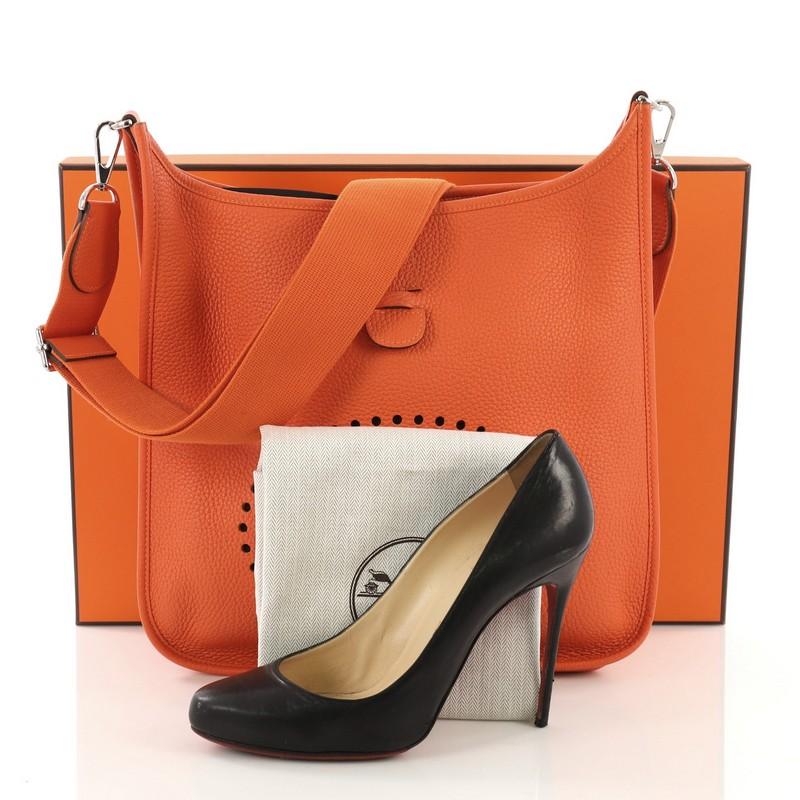 This Hermes Evelyne Crossbody Gen III Clemence GM, crafted in Feu orange Clemence leather, features signature perforated H design at the front, accessible back pocket, adjustable textile shoulder strap, and palladium hardware. It opens to a Feu