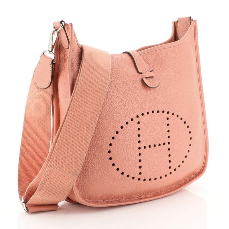 This Hermes Evelyne Crossbody Gen III Clemence PM, crafted in Crevette pink Clemence leather, features perforated H design at the front, accessible back pocket, adjustable textile shoulder strap, and palladium hardware. It opens to a Crevette pink