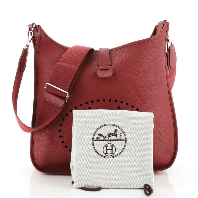 This Hermes Evelyne Crossbody Gen III Togo PM, crafted in Rouge Garance red Togo leather, features perforated H design at the front, accessible back pocket, adjustable textile shoulder strap, and palladium hardware. It opens to a Rouge Garance red