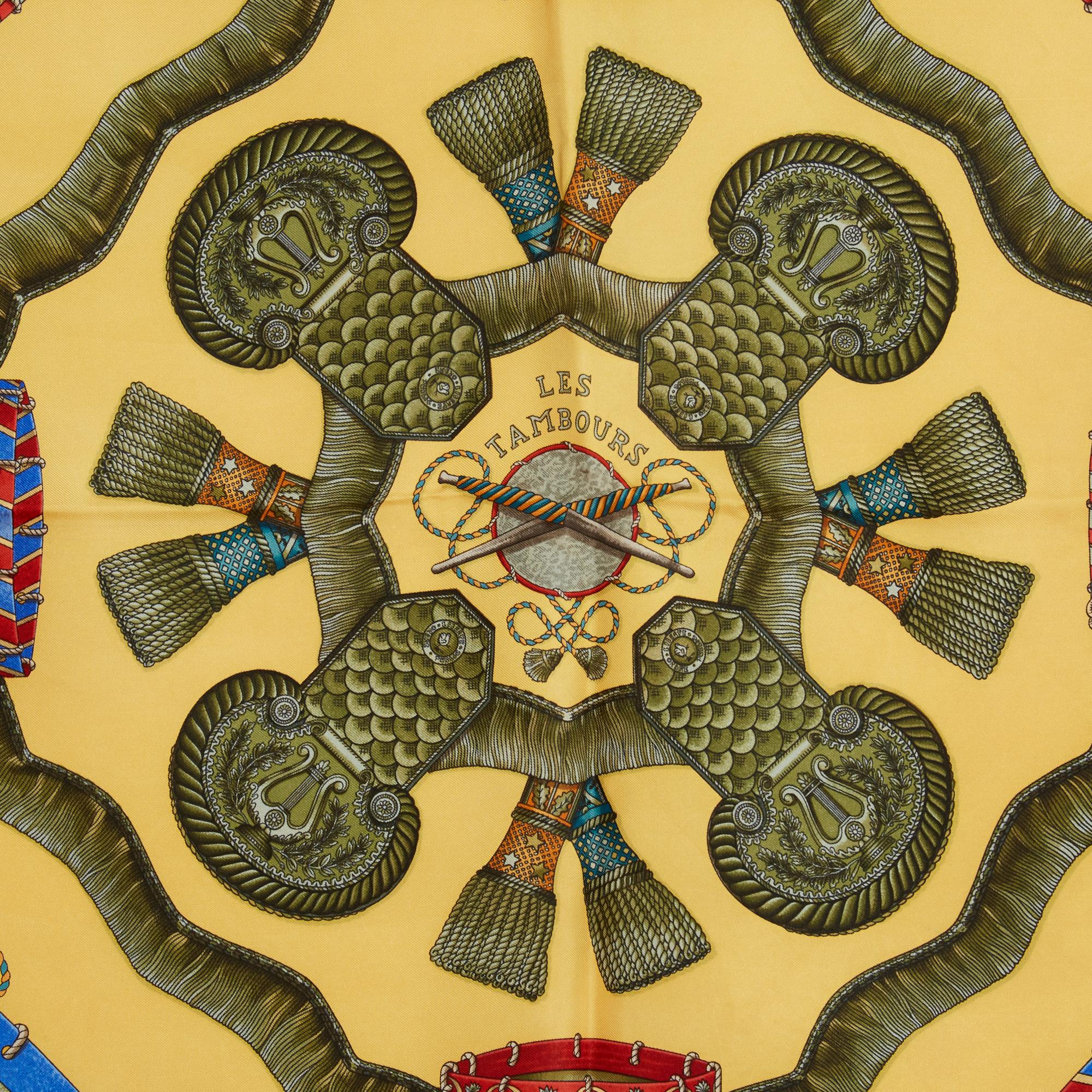 Pañuelo cuadrado Hermès 90 de sarga de seda, motivo Les Tambours de Joachim Metz, publicado en 1989 y reeditado en 1990, fondo amarillo pálido, bordes amarillo anaranjado. Ancho 90 cm x largo 90 cm aproximadamente. El pañuelo es vintage y