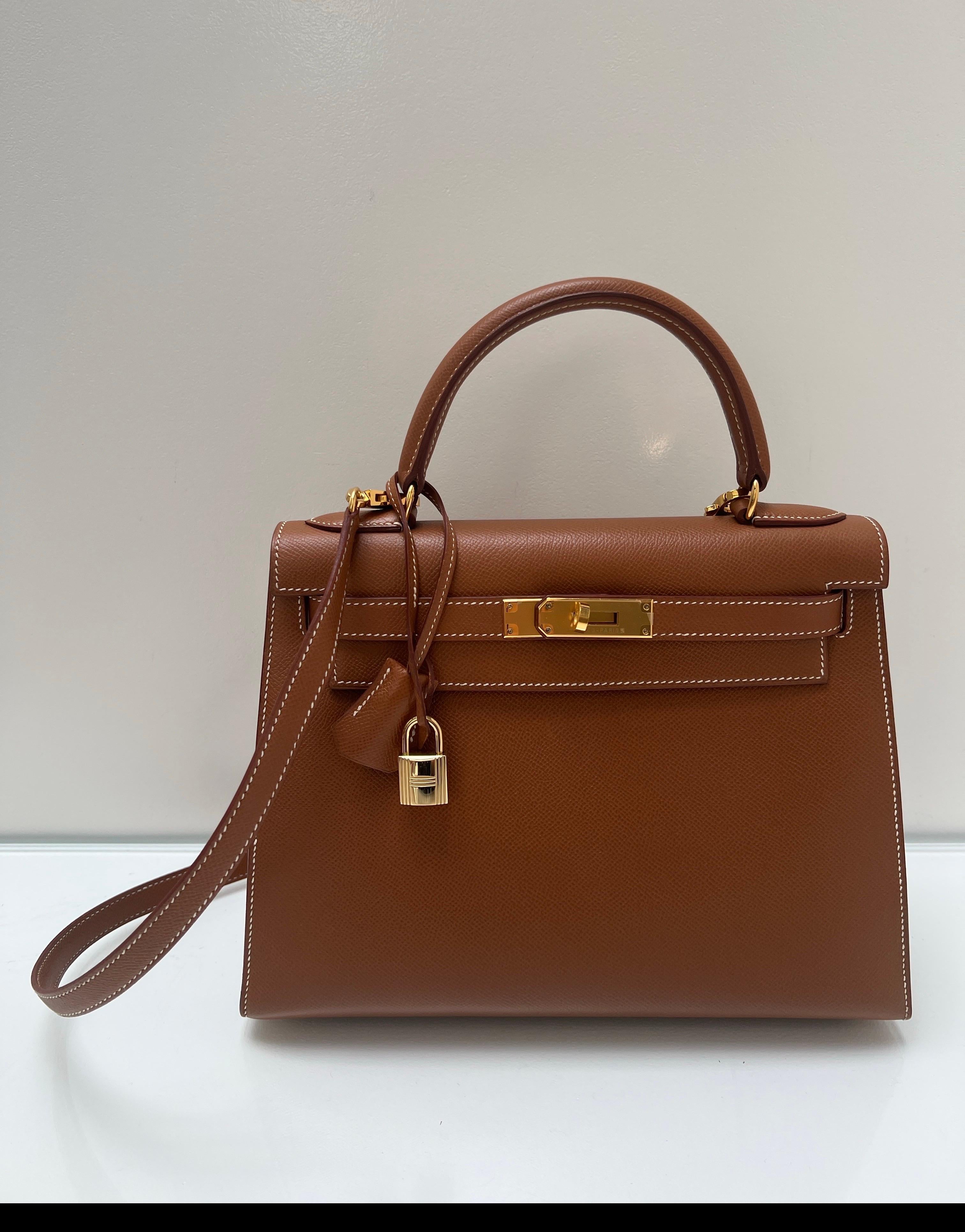 Borsa Kelly 28 in oro di Hermes. Colore oro e marrone con finiture dorate. Sellier Kelly ha una struttura. Cuciture a contrasto bianche. Interni puliti. Plastica sulla ferramenta. Sembra nuovo, in ottime condizioni. Include cinturino, clochette,