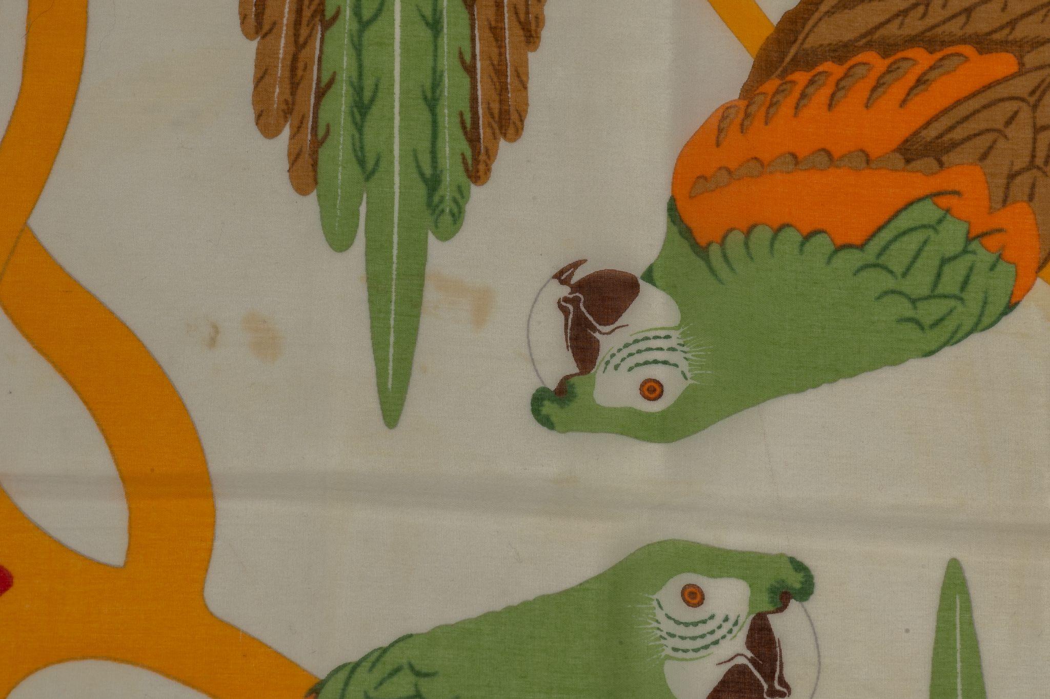 Hermès Green Parrots Cotton Sarong. The pattern features several parrots in the color green, brown and orange. The frame is red and has rolled edges. Piece is in very good condition with a stain. Please see pictures.