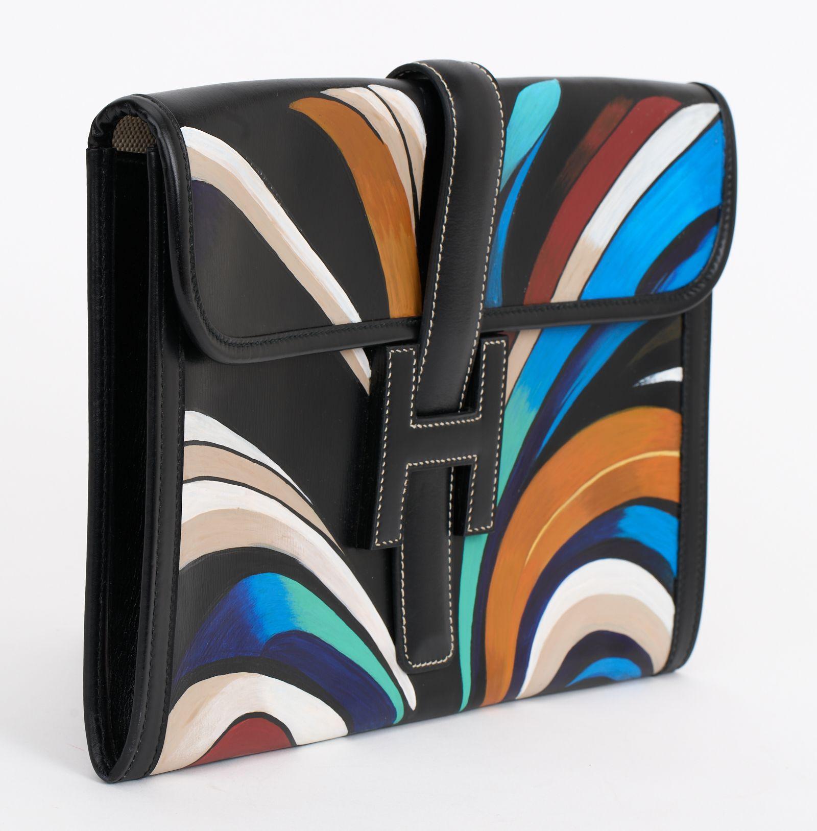 The Hermès Hand Painted Jige Clutch in black box calf leather features an art deco hand painted design, inspired by the Chrysler building in New York. Front strap flap H closure opens to a woven canvas lined interior. Clean corners and edges. The