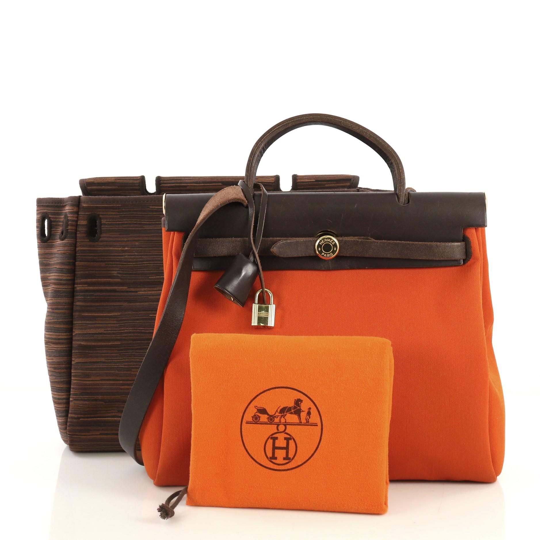 This Hermes Herbag Vibrato and Leather PM, crafted from Ebene brown Vache Hunter leather and Ebene brown and Orange H orange Vibrato, features leather top handle, long flat leather shoulder strap, top frame, and gold hardware. Its leather strap