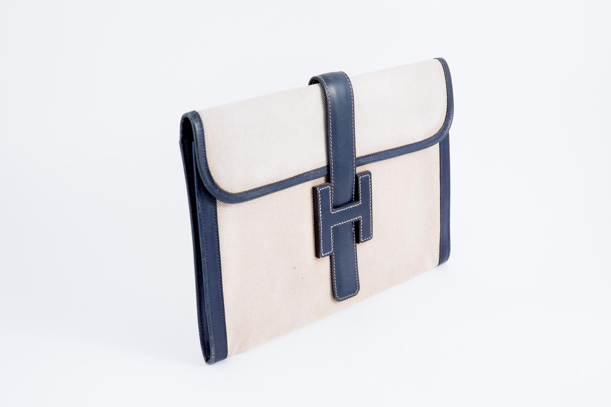 Hermes Jigé ivory canvas and navy box leather clutch featuring a foldover top, a strap closure, a front logo patch, a cotton canvas lining, a center front Hermes Paris stamp. 
In good vintage condition. Made in France.
Width:11.4in. (29cm)
Height: