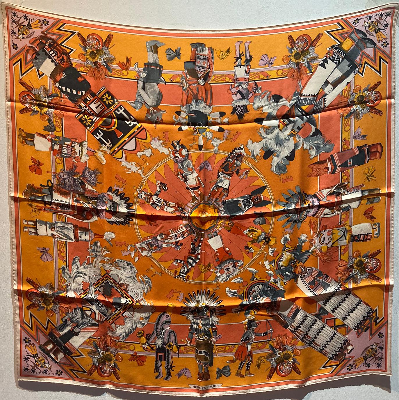 Hermes Kachinas Silk Scarf in excellent condition. Original silk screen design c1992 by Kermit Oliver features various multicolor native american chiefs and people in traditional dress over an orange background. This is a highly collectible piece as
